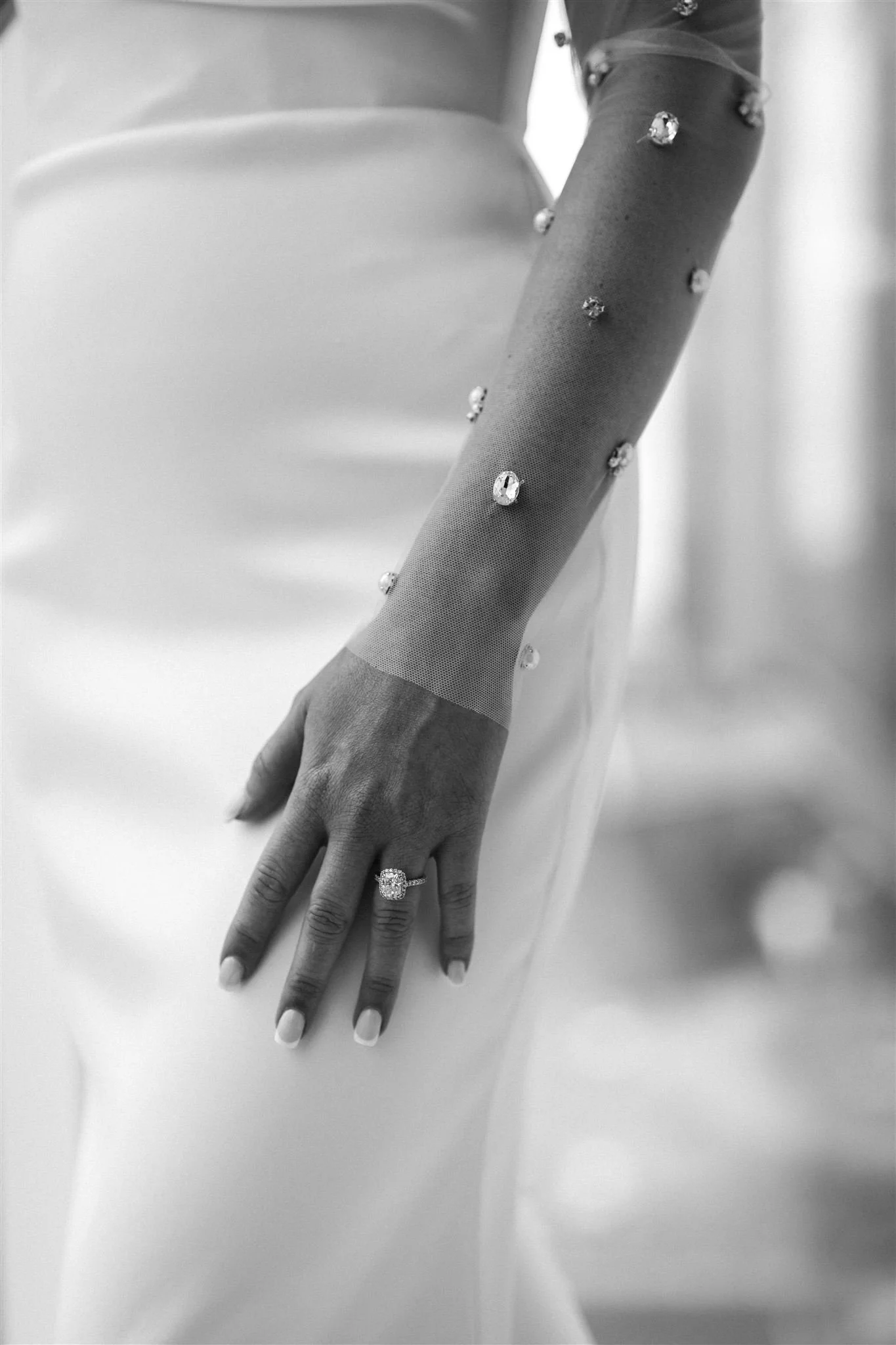 Close-up of a woman's hand resting on her thigh, showing a large engagement ring, and her arm adorned with a sheer, embellished glove with rhinestones or crystals.