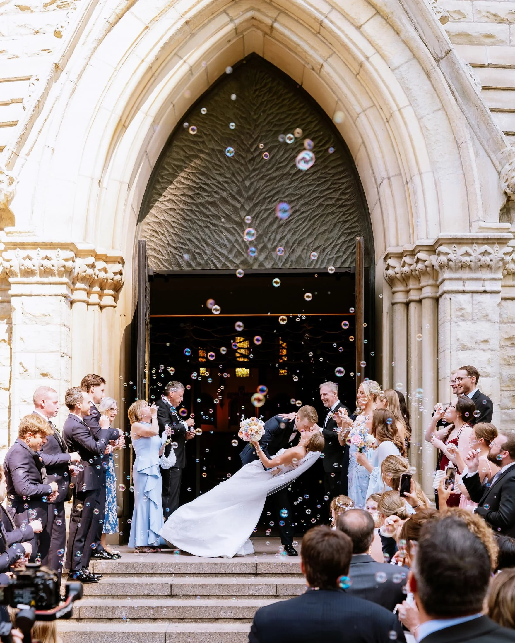 A newlywed couple kissing on the steps of a church with friends and family around celebrating their wedding, surrounded by soap bubbles.