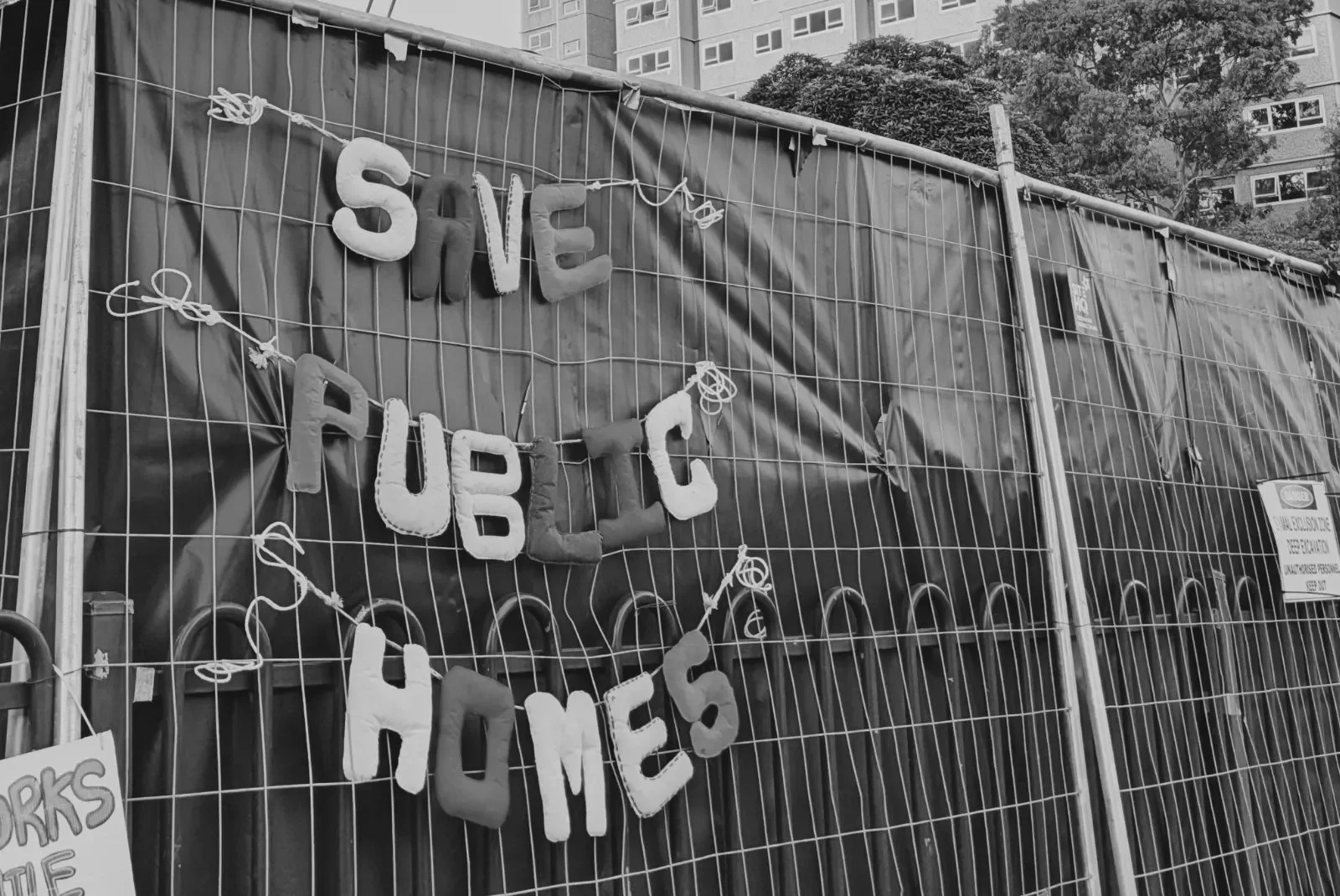A black and white photo of a construction fence with a sign that says "Save Public Homes" spelled out with large letters made of foam or fabric, attached with string. There is a smaller sign on the fence that says "Works" and some trees and apartment buildings in the background.