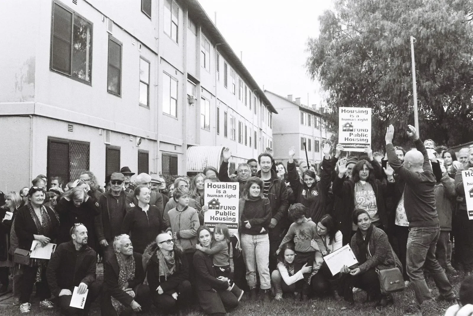 A large crowd of people gathered at a protest march. Some are holding signs that read 'Housing is a human right' and 'Fund Public Housing.' The crowd appears to be diverse, with people of different ages and ethnicities, standing and sitting in front of a residential building with multiple windows, during the daytime.