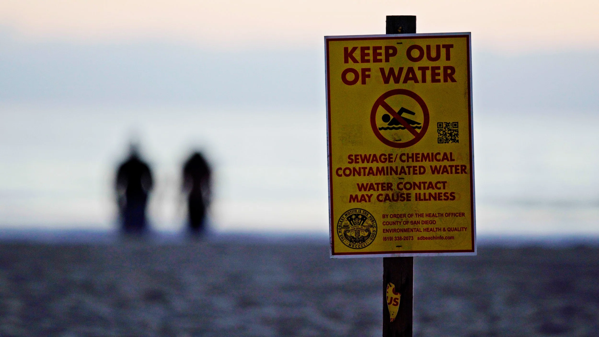 A sign on a beach with two people in the background.
