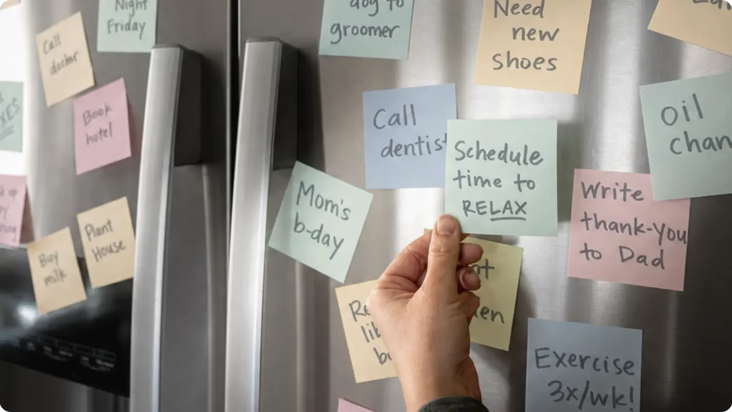 A hand reaches for a sticky note reading 'Schedule time to relax' among dozens of task reminders covering a fridge door.