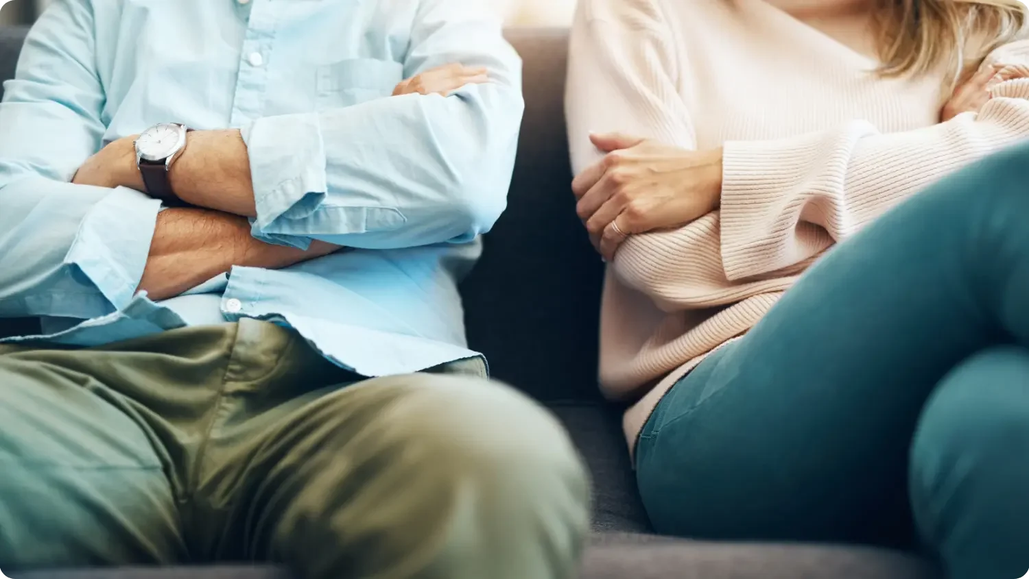 A man and a woman sit apart on a sofa with their arms folded, turned slightly away from each other in tense silence