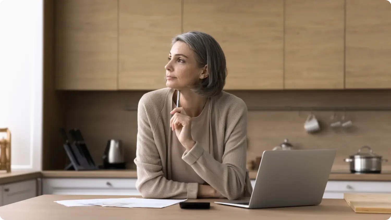 A woman rests on a kitchen counter with a pen to her chin, looking thoughtfully into the distance beside her laptop and paperwork.