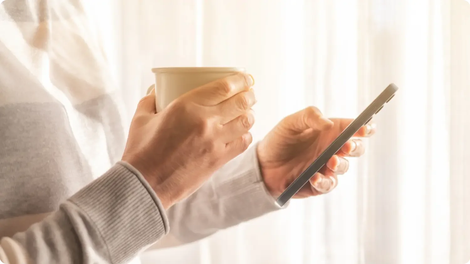 A person in a soft grey jumper holding a coffee cup in one hand and checking their phone in the other, by a sunlit window.