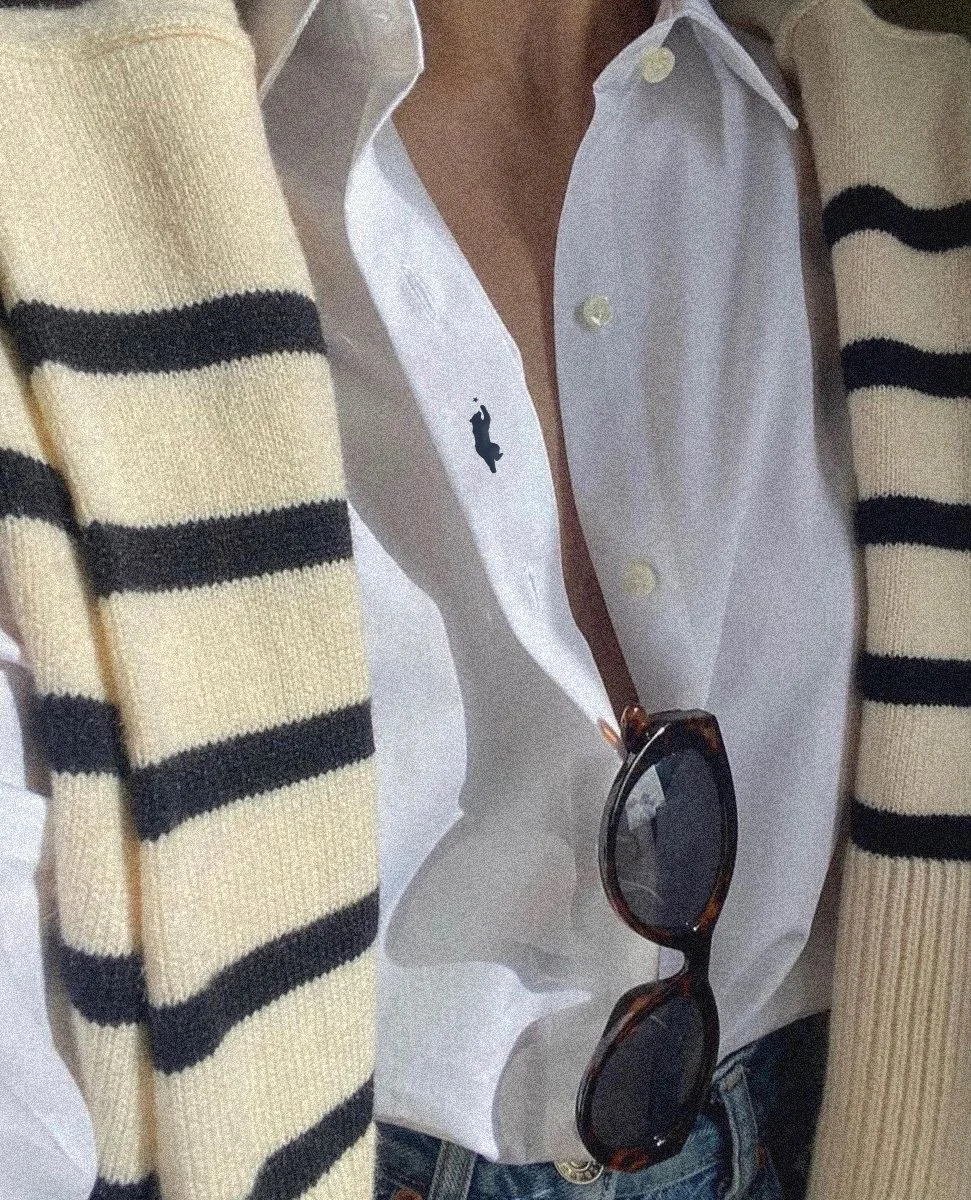 Une personne porte une chemise blanche avec un logo de cheval noir, un pull à rayures beige avec des bandes noires, et des lunettes de soleil avec une monture marron.