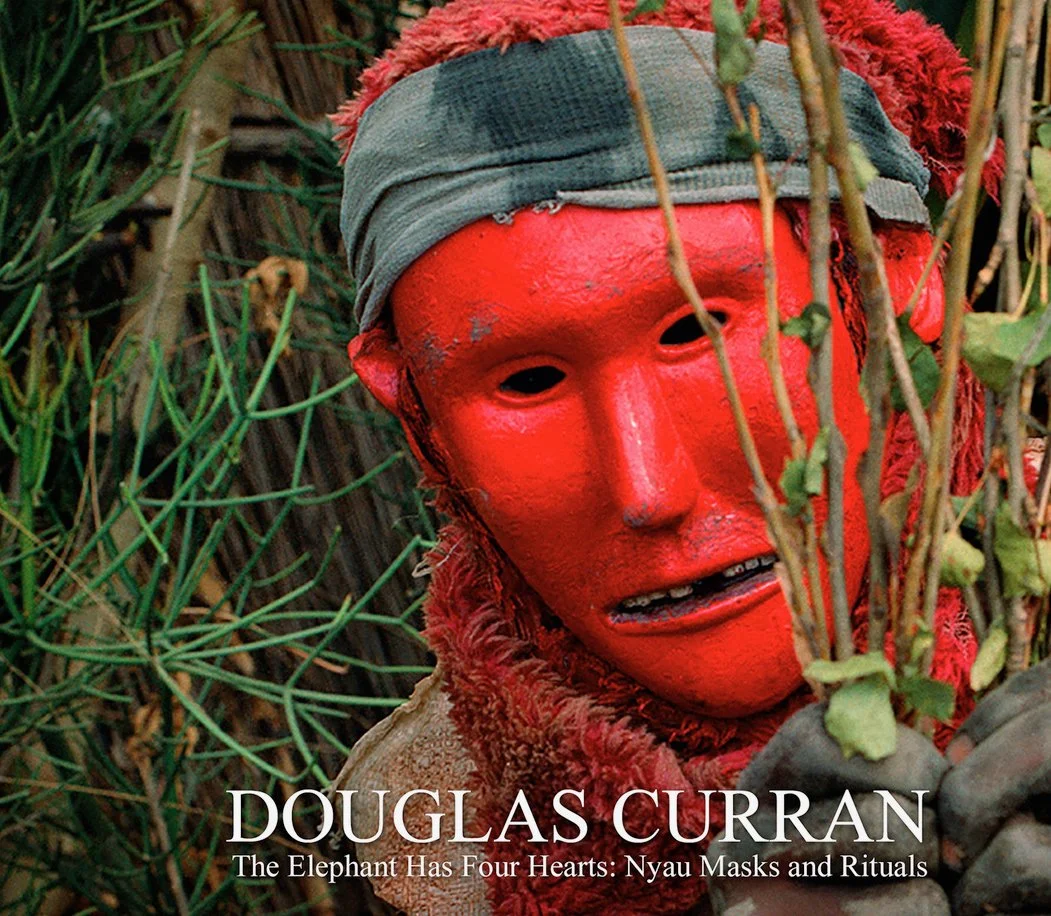 A masked spirit dancer in red mask peers through a bundle of green branches.