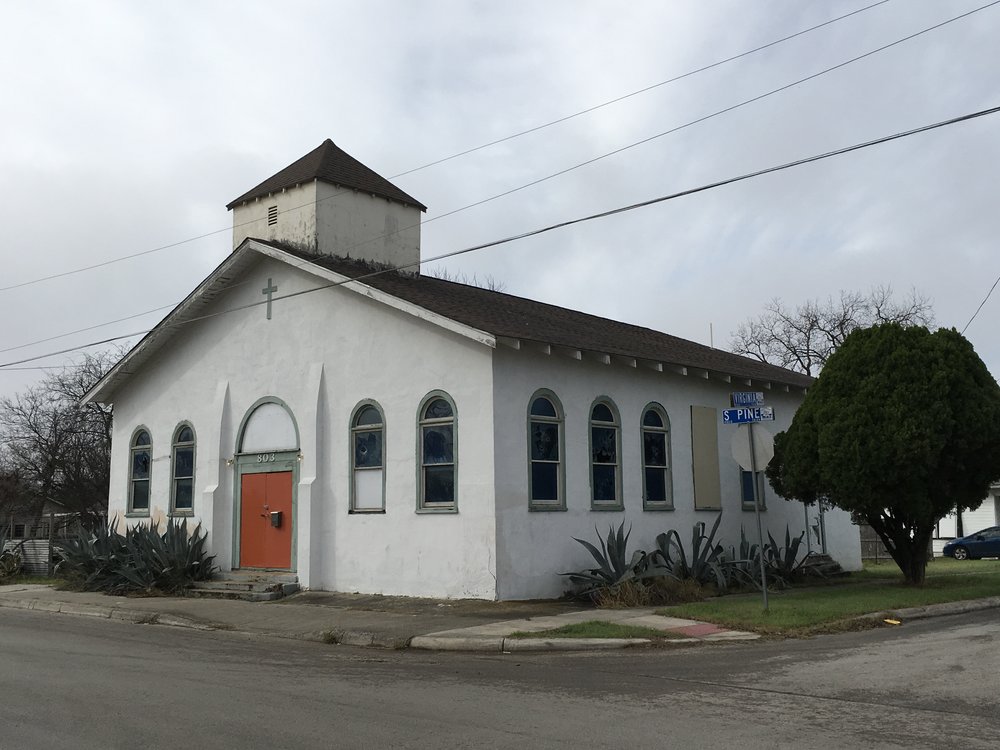  White stucco church with front gable roof, arched wooden windows, an orange front door with green trim with a green cross hung above it, and a squat square steeple. 