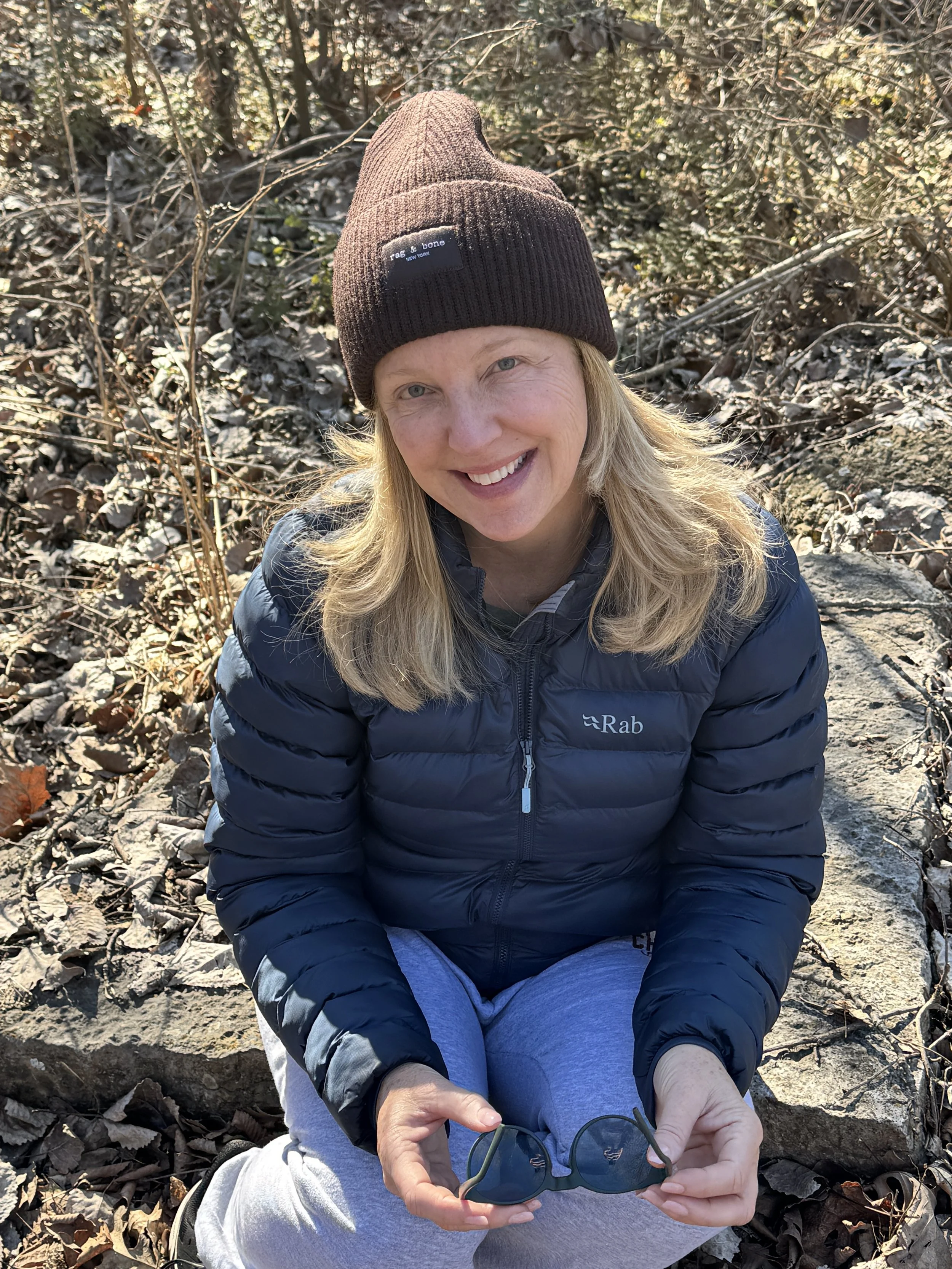 A woman with blonde hair, wearing a brown knit beanie and a blue puffy jacket, sitting outdoors among fallen leaves, holding sunglasses, with sunlight shining on her face.