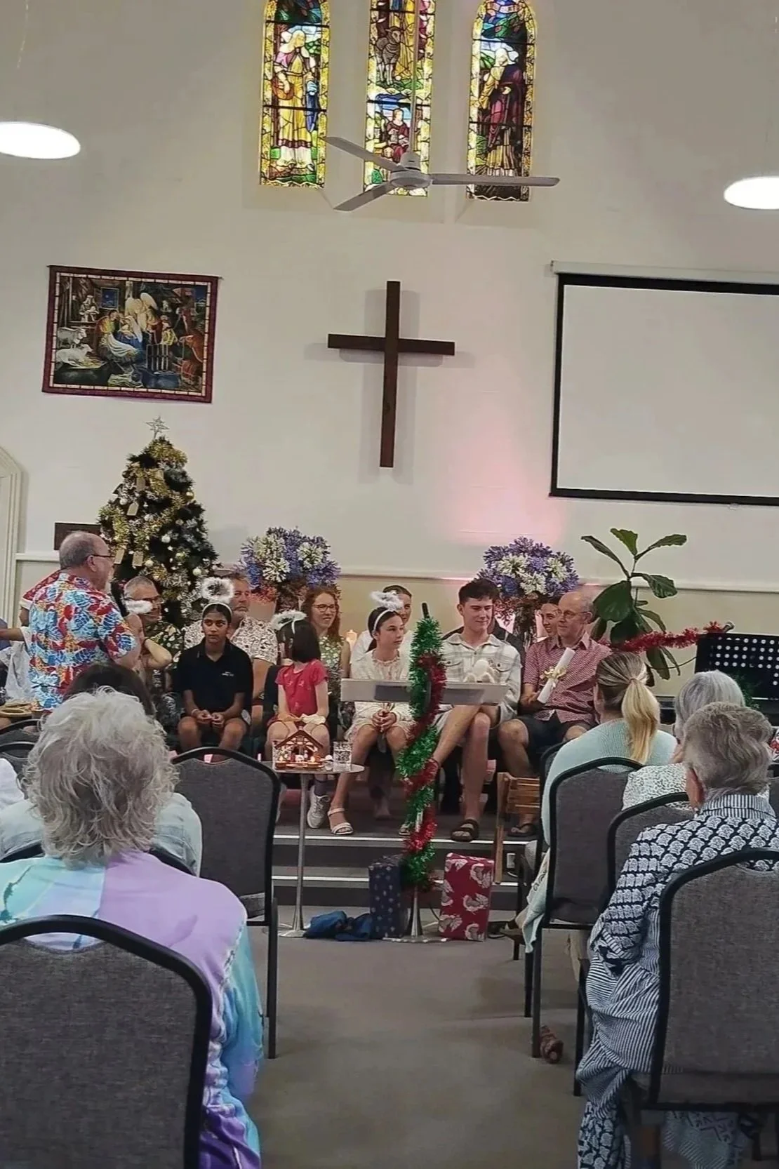 People gathered in a church for a Christmas celebration, with a Christmas tree decorated with gold and white ornaments, wrapped gifts underneath, and children seated on stage, some holding Christmas-themed props. The church features stained glass windows, a large wooden cross, and religious artwork on the walls.