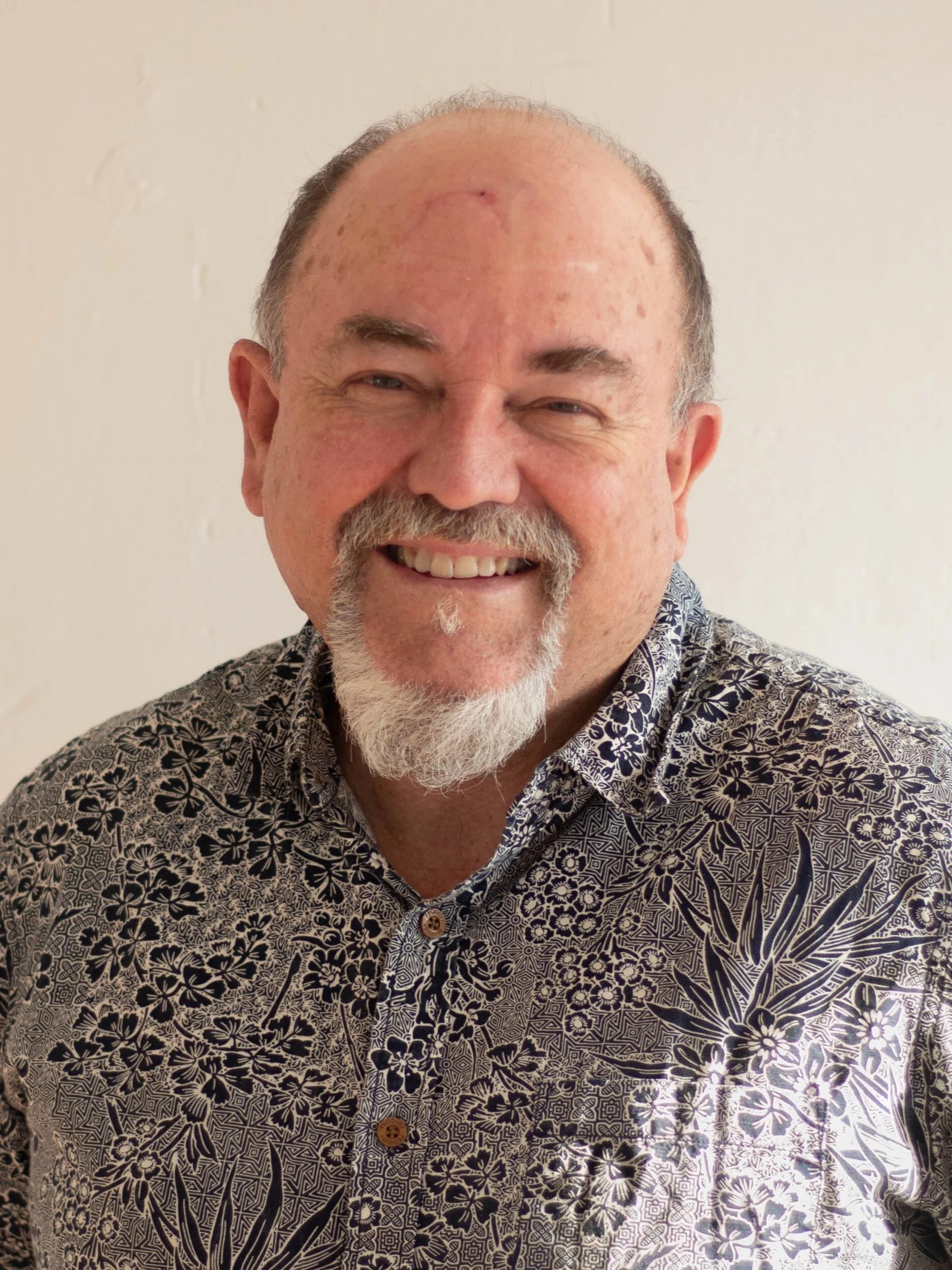 A smiling middle-aged man with a beard and mustache, wearing a patterned button-up shirt, standing in front of a plain wall.