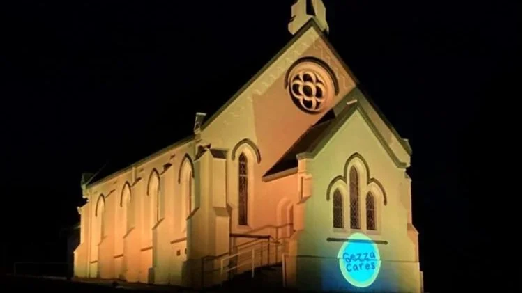 A church illuminated by colored lights at night, with a blue sticker that reads 'Fezza Cares' on the front.