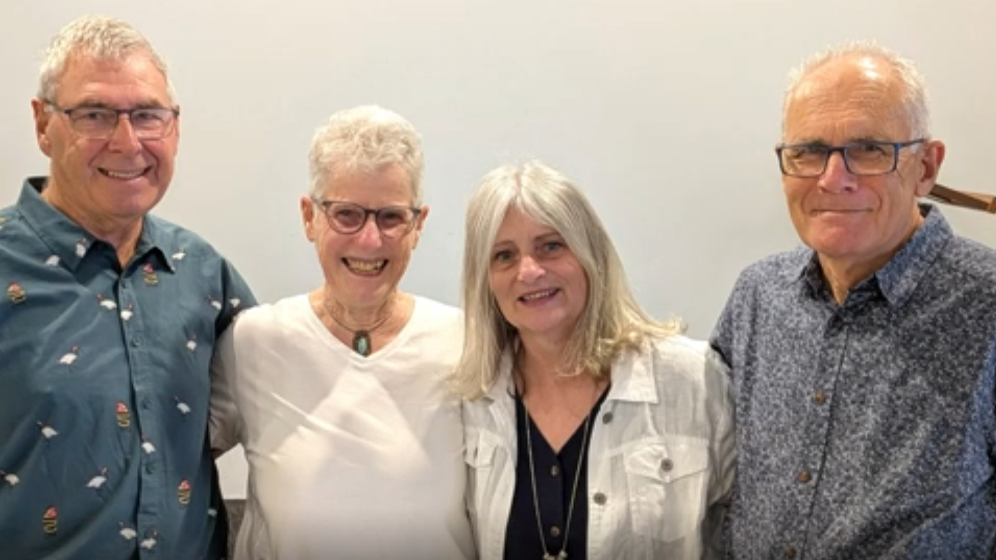 Four people standing together, smiling, in an indoor setting against a plain background.