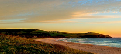Sunset over a beach with grassy dunes and a rocky headland in the distance.