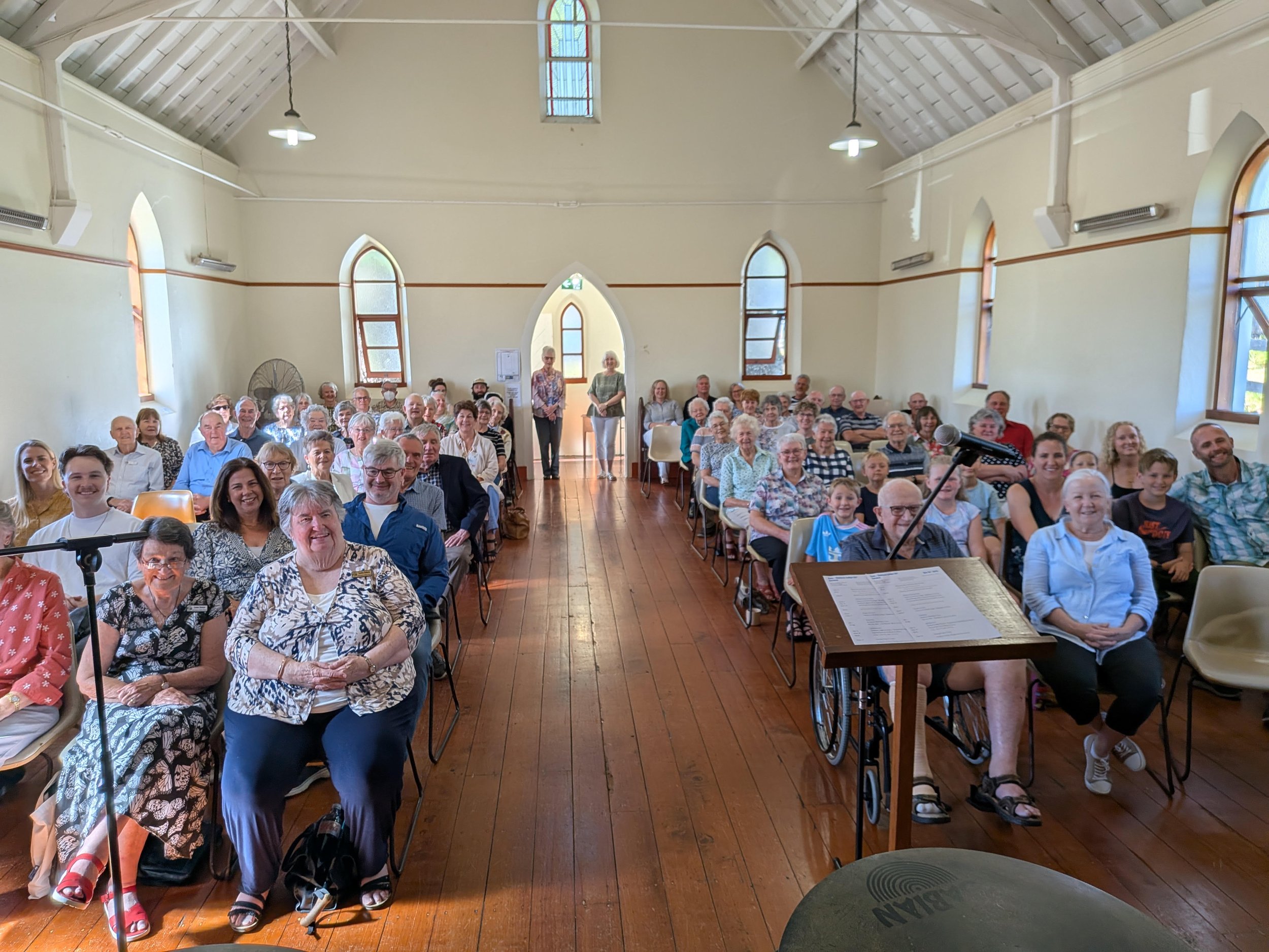 An indoor gathering of mostly elderly men and women, with some younger individuals, seated and smiling in a church or community hall with wooden floors and high arched windows; a music stand and microphone are visible in the foreground.