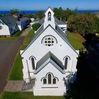 Aerial view of a white church building with a steeple, surrounded by green lawns and trees, with a body of water in the background.