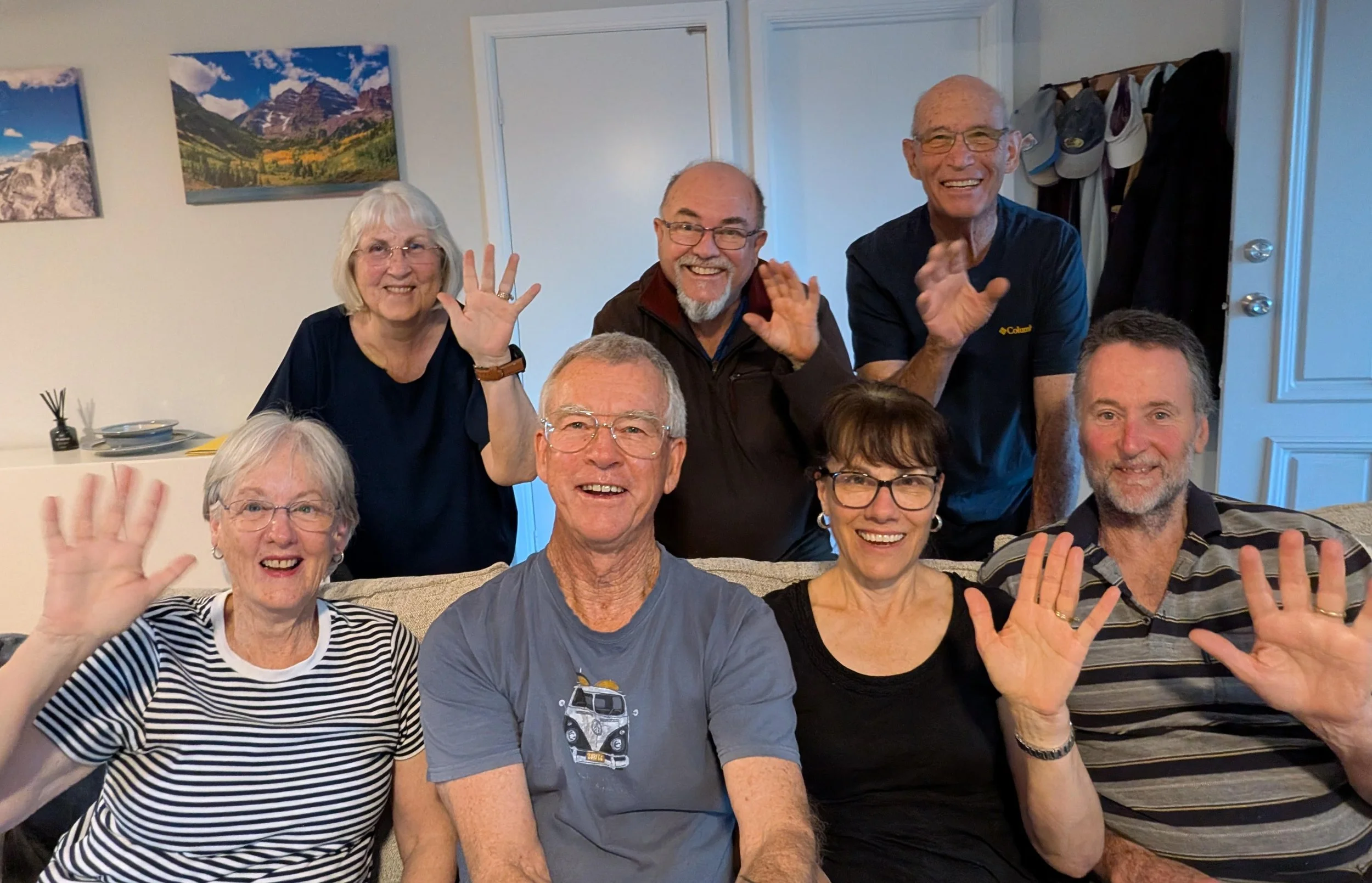 Group of seven smiling adults waving at the camera in a living room