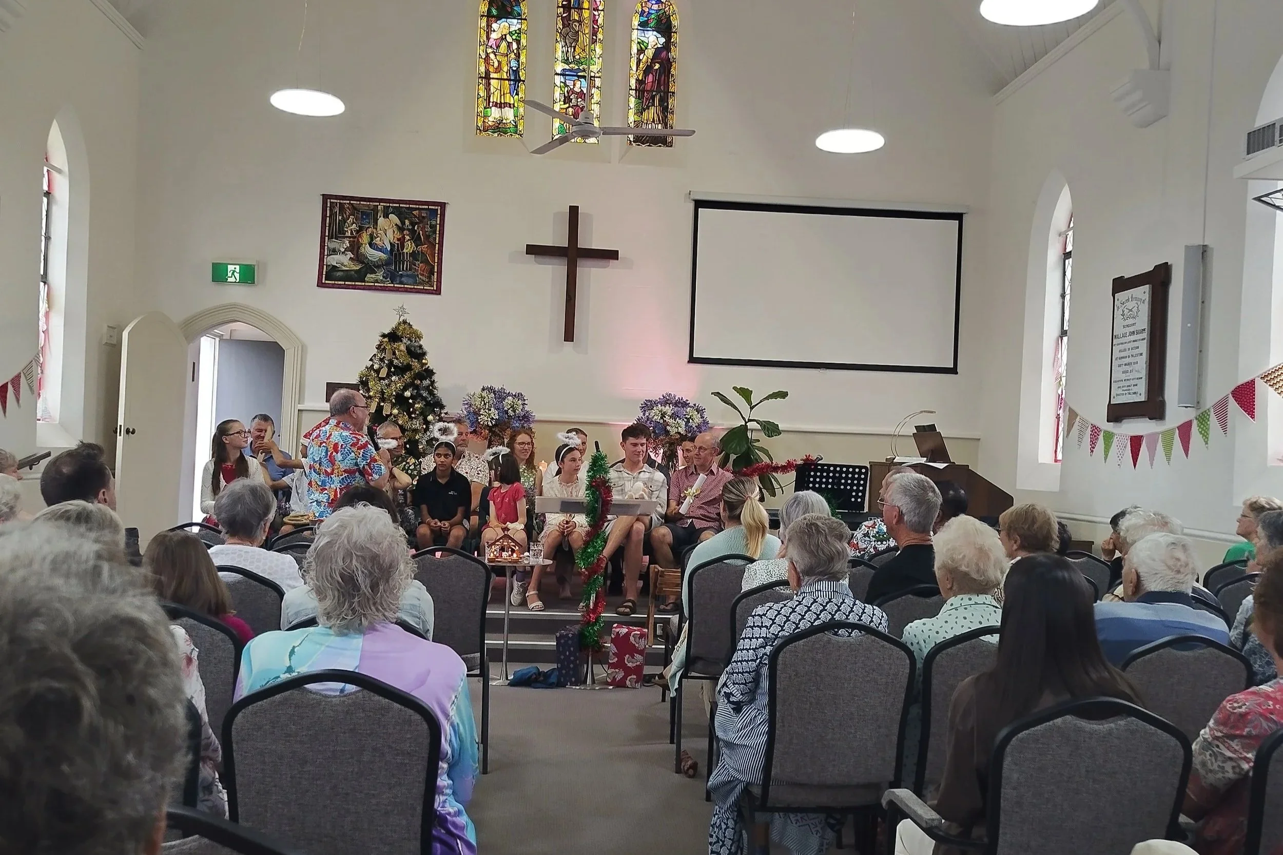 A church decorated for Christmas with a Christmas tree, flowers, and festive decorations. People are seated facing a stage where a group of children and adults are gathered, some holding musical instruments and papers, with a man in a colorful shirt speaking. The church has stained glass windows, a cross on the wall, and an audience of elderly and younger individuals.