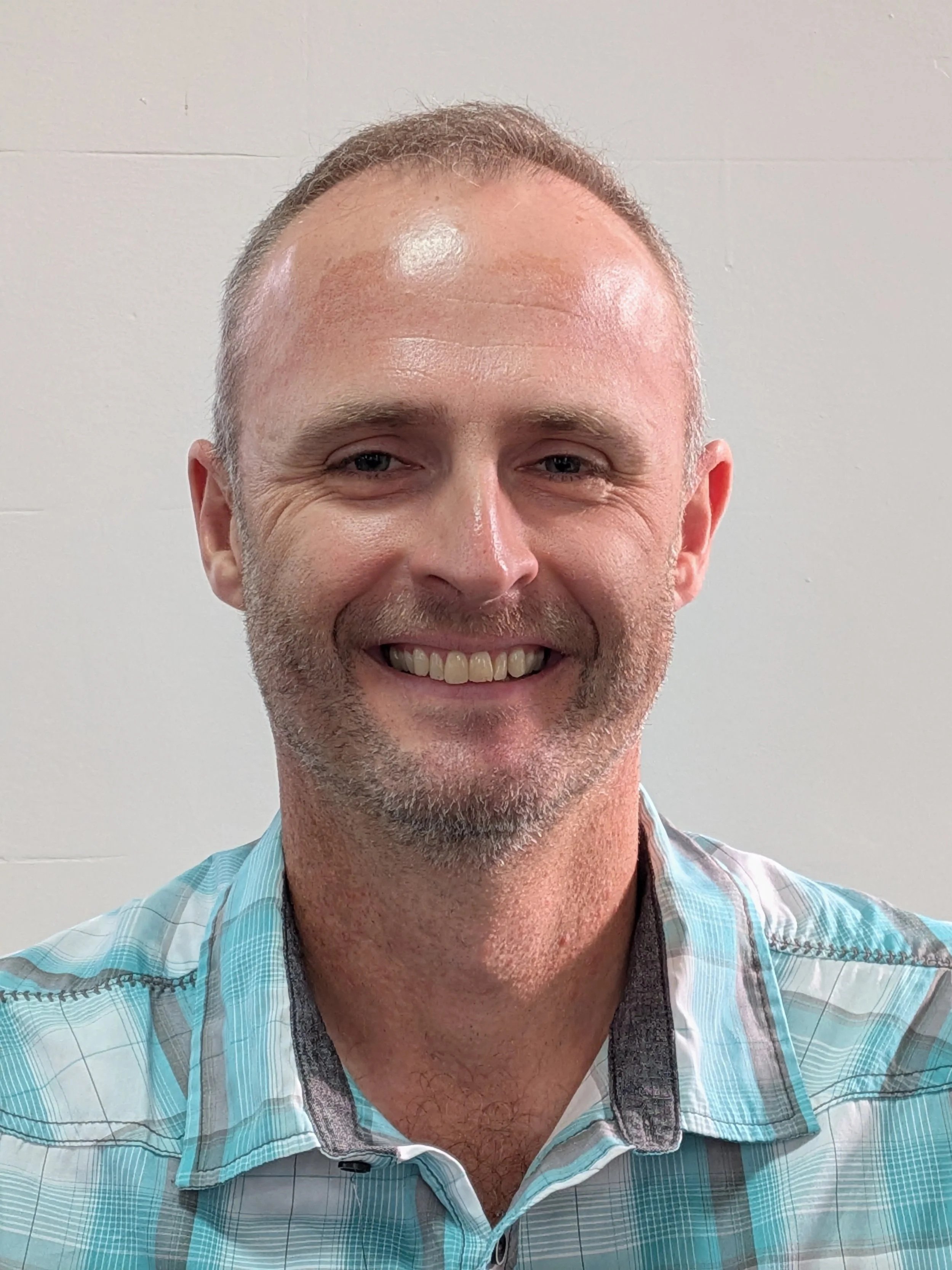 A smiling man with short light-colored hair, a beard, wearing a blue plaid shirt, standing in front of a plain white wall.