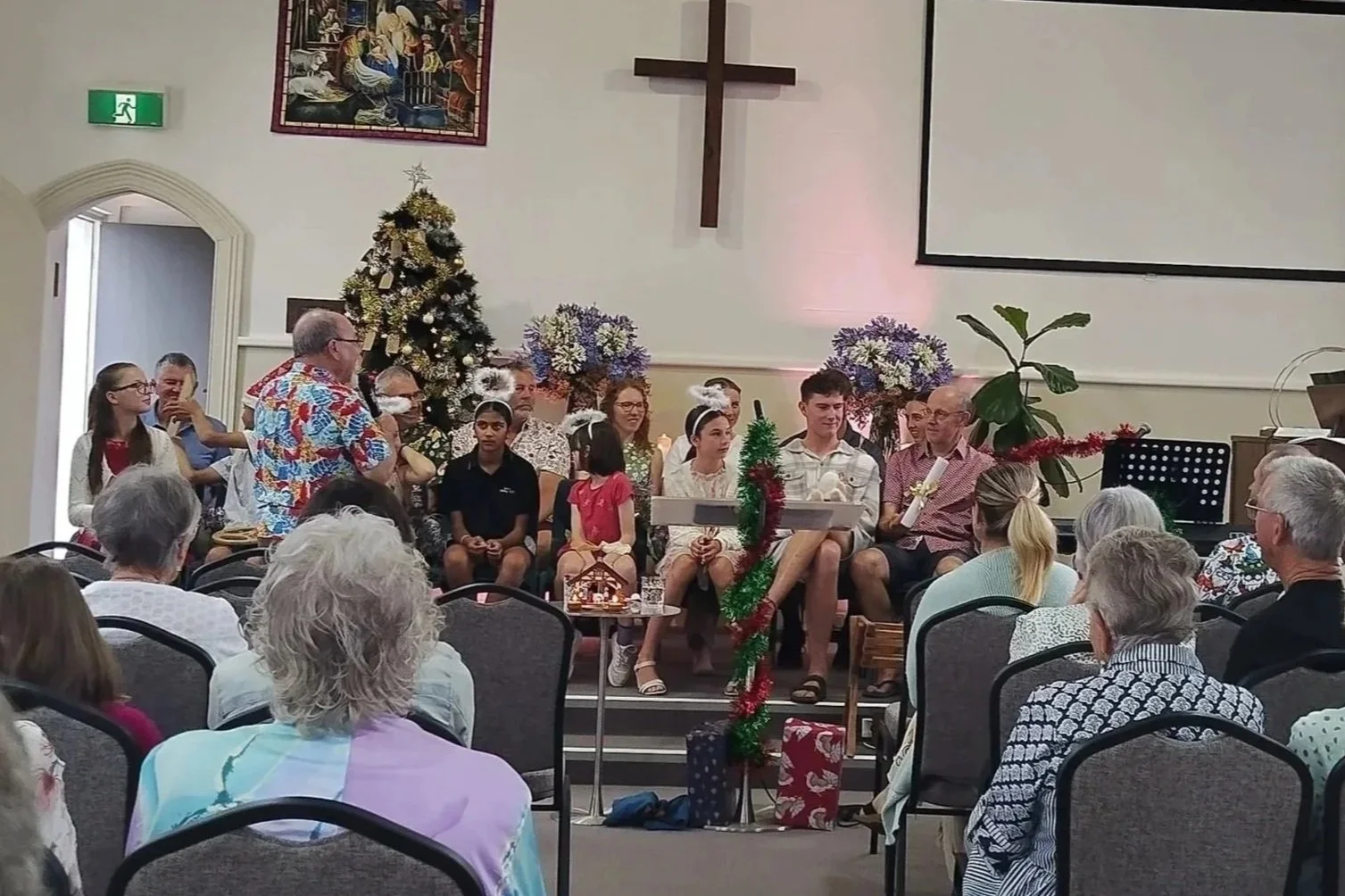 A Christmas event in a church with a decorated Christmas tree, a cross on the wall, and people sitting on a stage, including children and adults, some holding gifts and wearing festive attire. Audience members are seated facing the stage.