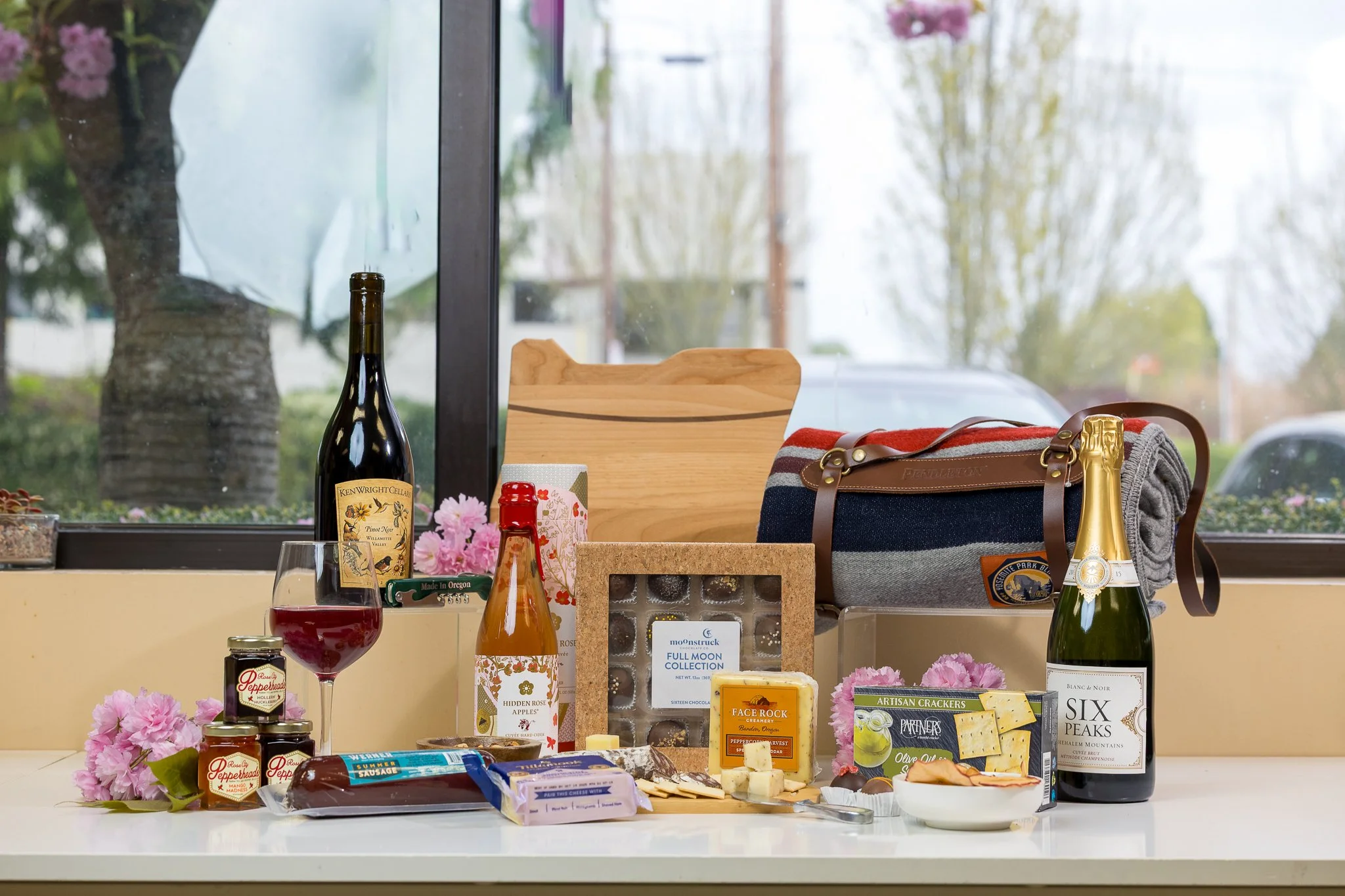 A display of various food and drink products on a white table, including wine bottles, jars, cheese, crackers, and chocolates, with pink flowers and a window reflecting trees outside in the background.