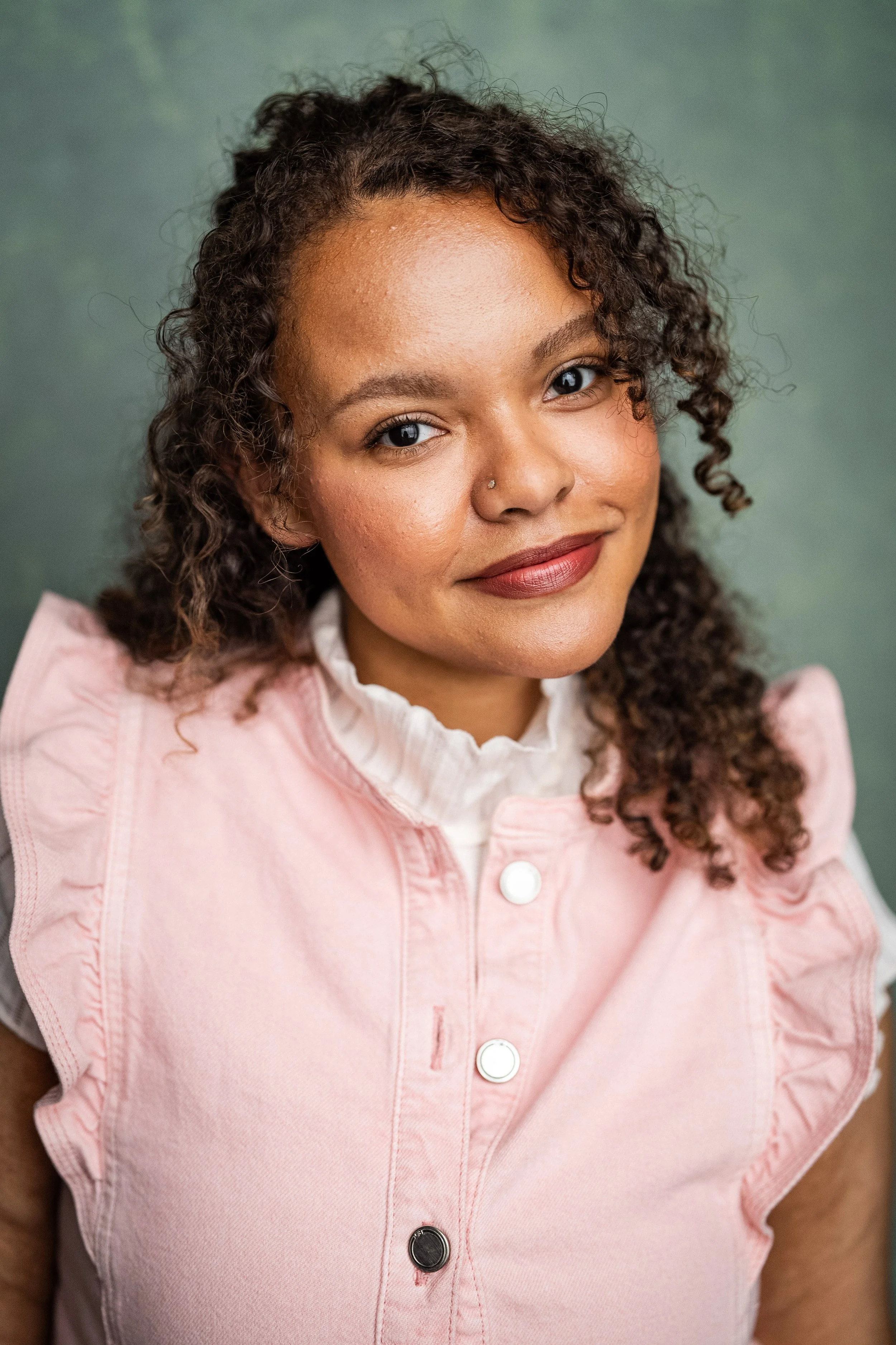 Headshot of Lydia-Renee Darling.  A young woman with curly hair, wearing a pink vest over a white ruffled shirt, smiling.