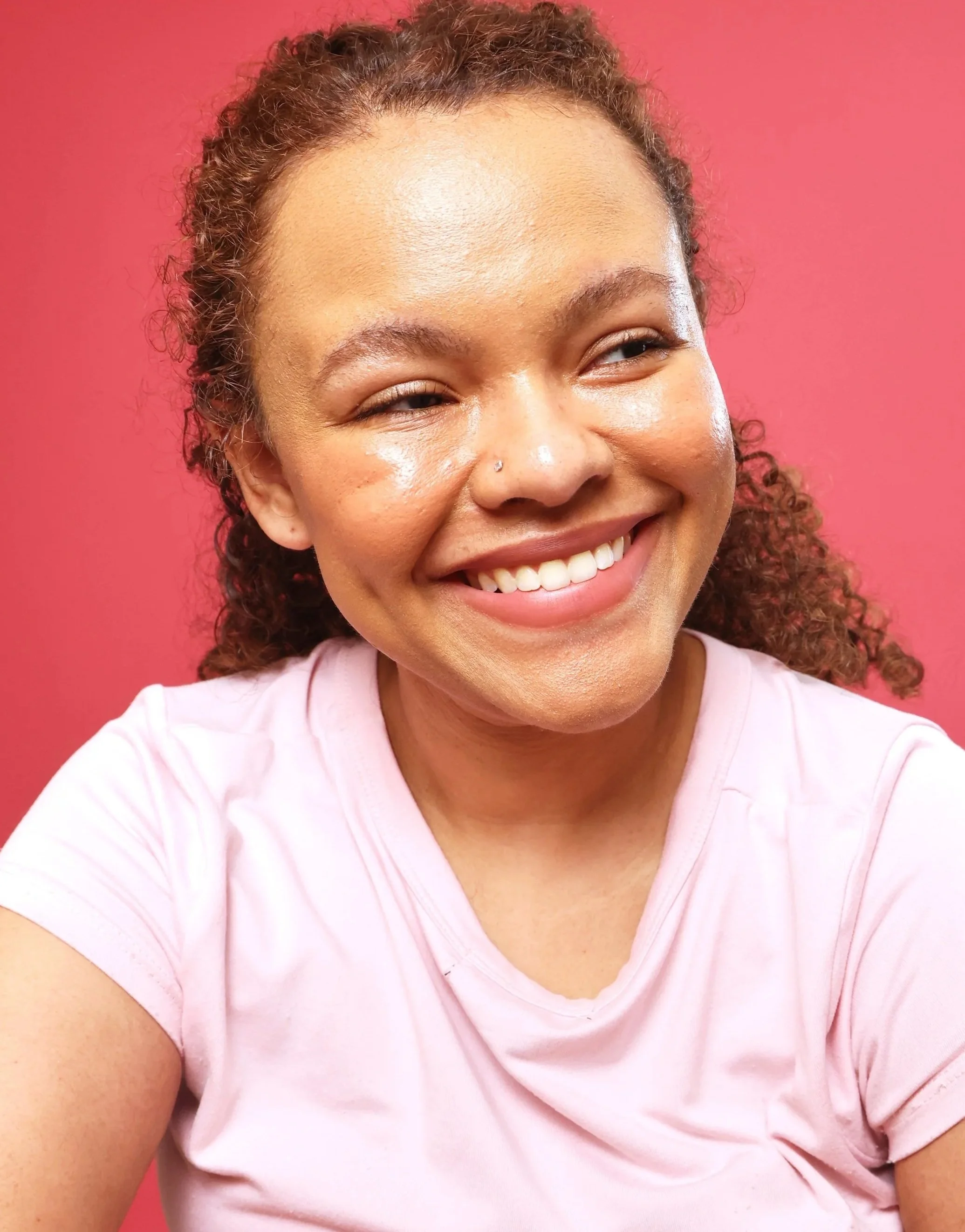 Close-up of a smiling woman with curly hair, wearing a light pink t-shirt, sitting against a pink background.