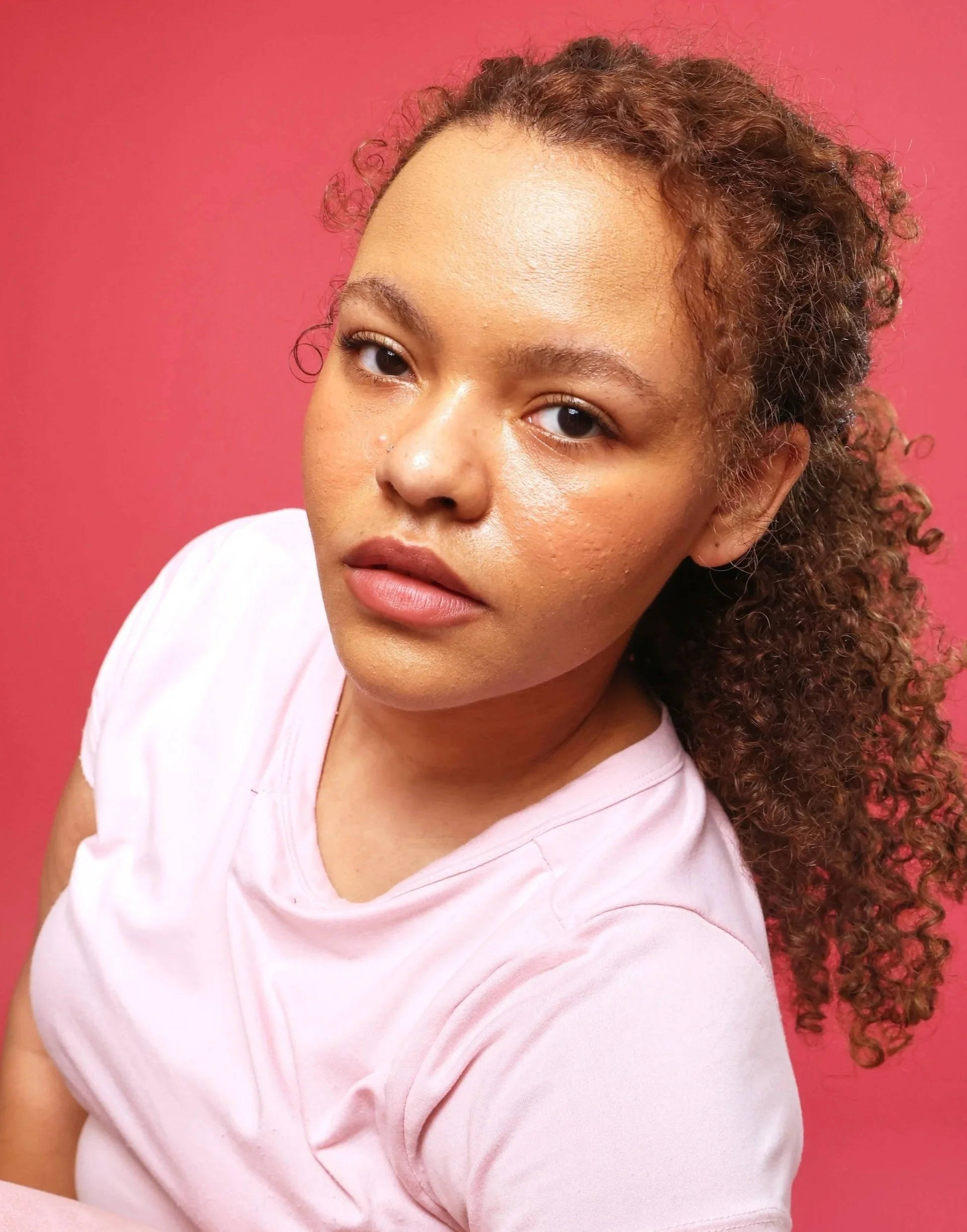 Close-up of a woman with curly brown hair and natural makeup, wearing a light pink T-shirt, against a pink background.