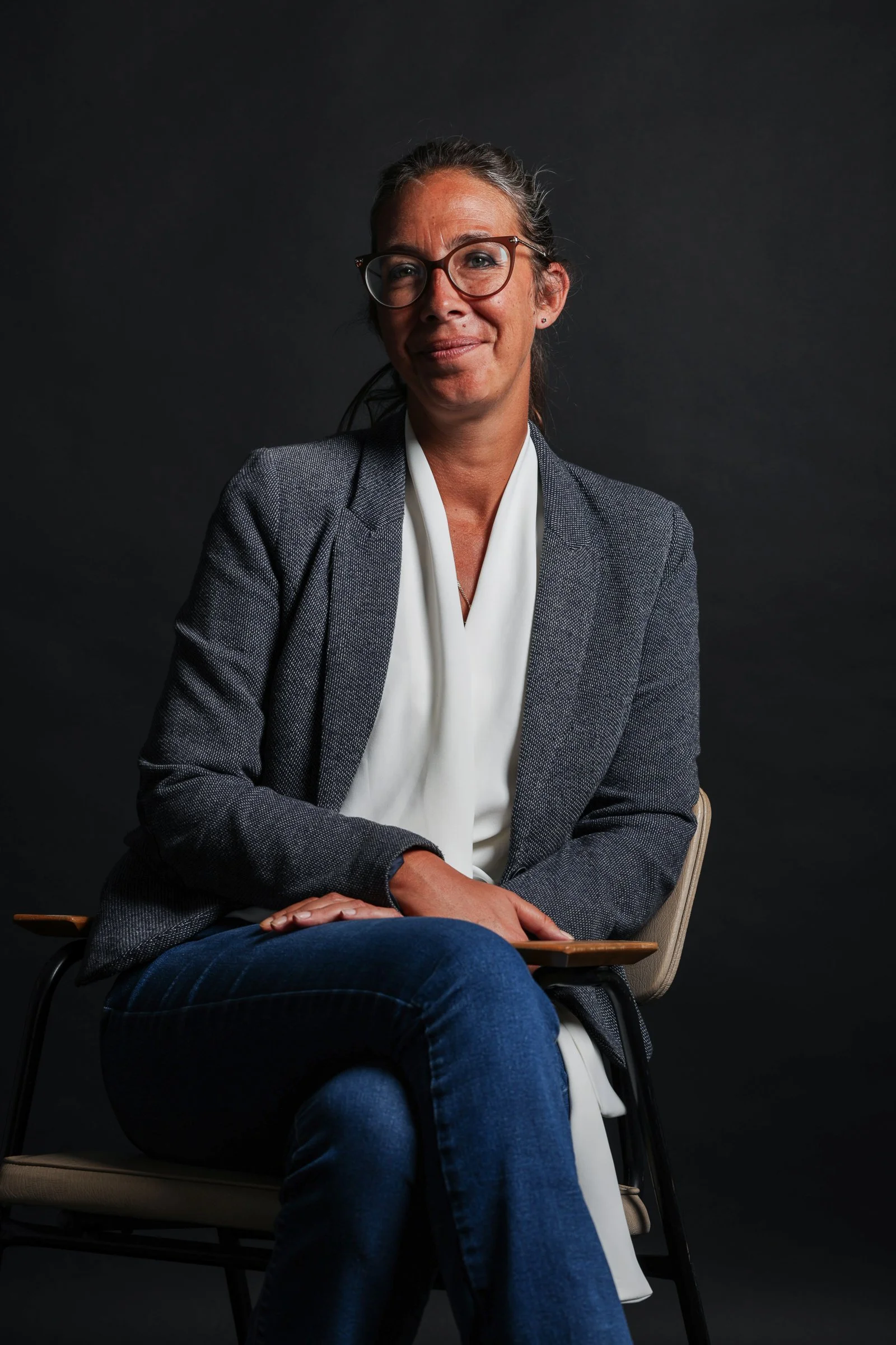 Une femme assise sur une chaise en bois, portant une veste grise, une blouse blanche et des lunettes, avec un fond noir.