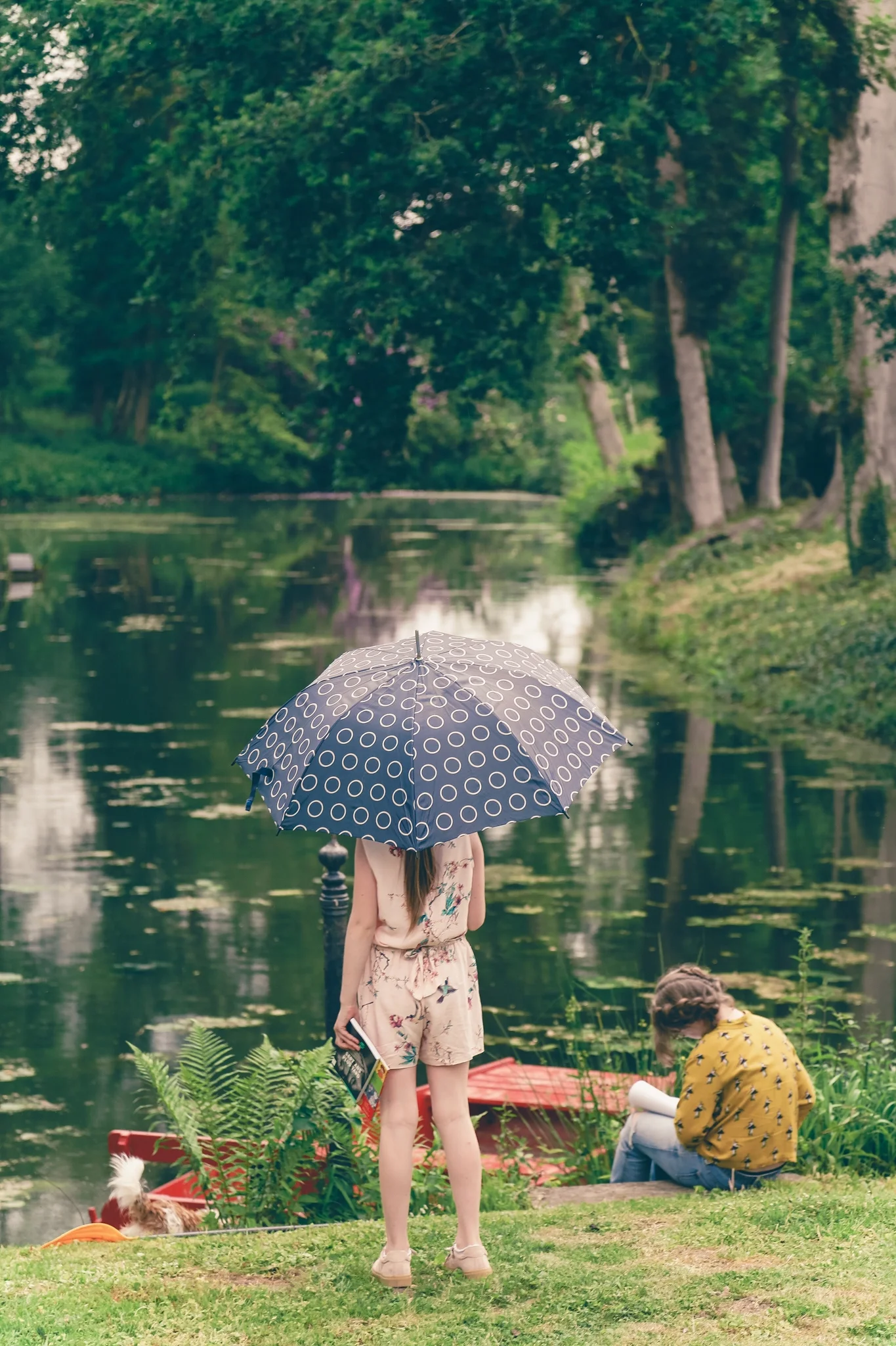 Deux jeunes filles près d'un lac, l'une tenant un parapluie noir à motifs blancs, l'autre assise sur l'herbe avec un carnet, en environnement verdoyant avec des arbres et un bateau rouge dans l'eau.