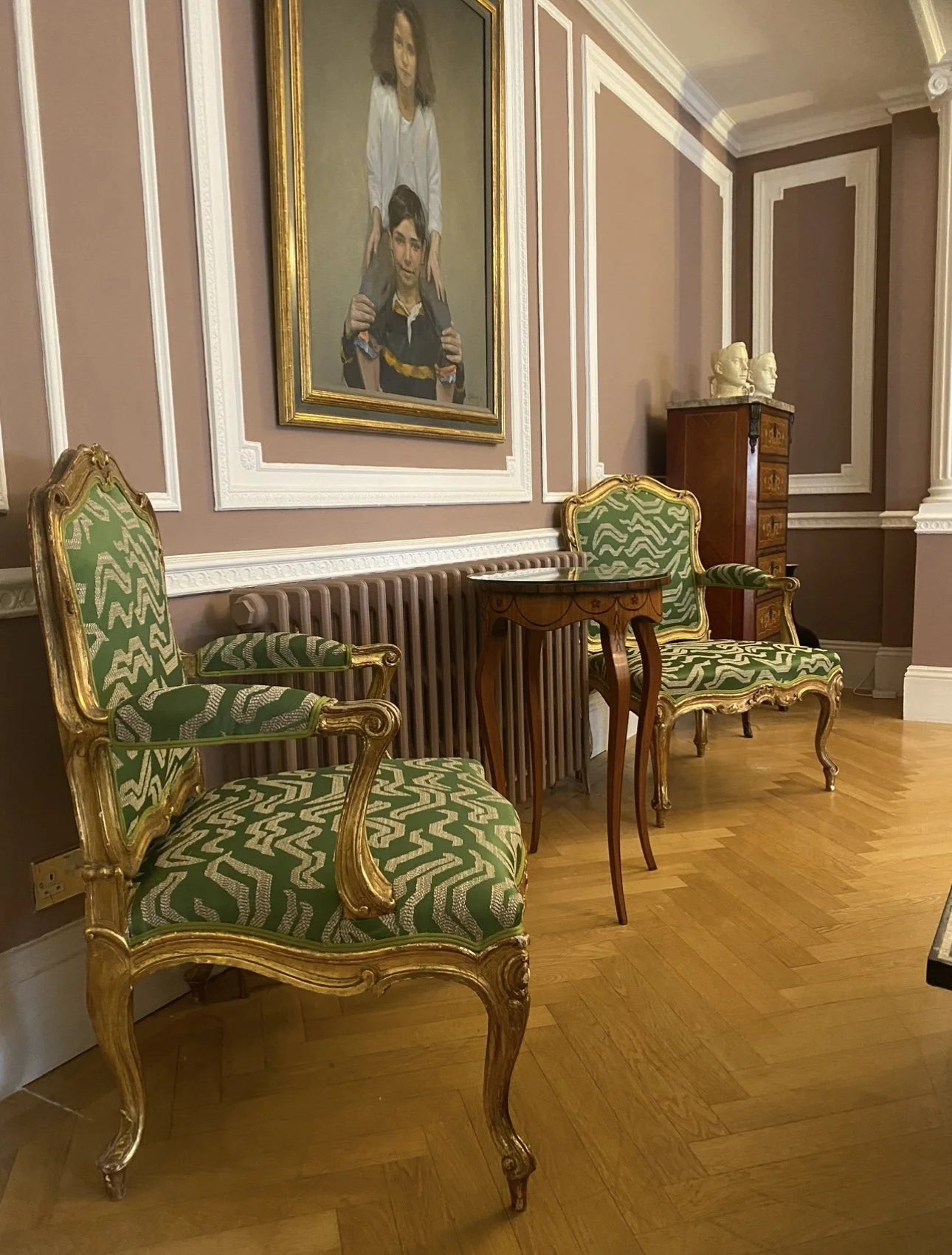 Elegant room with two green and gold patterned chairs, a small wooden table, a portrait painting, a wooden cabinet with bust sculptures, and a wooden floor.