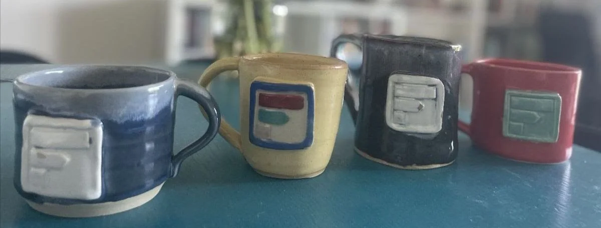 Four handmade ceramic mugs in blue, yellow, black, and red lined up on a teal surface. Each mug features a raised, rectangular emblem of a stylized bar graph, showcasing a rustic, artisanal glaze.