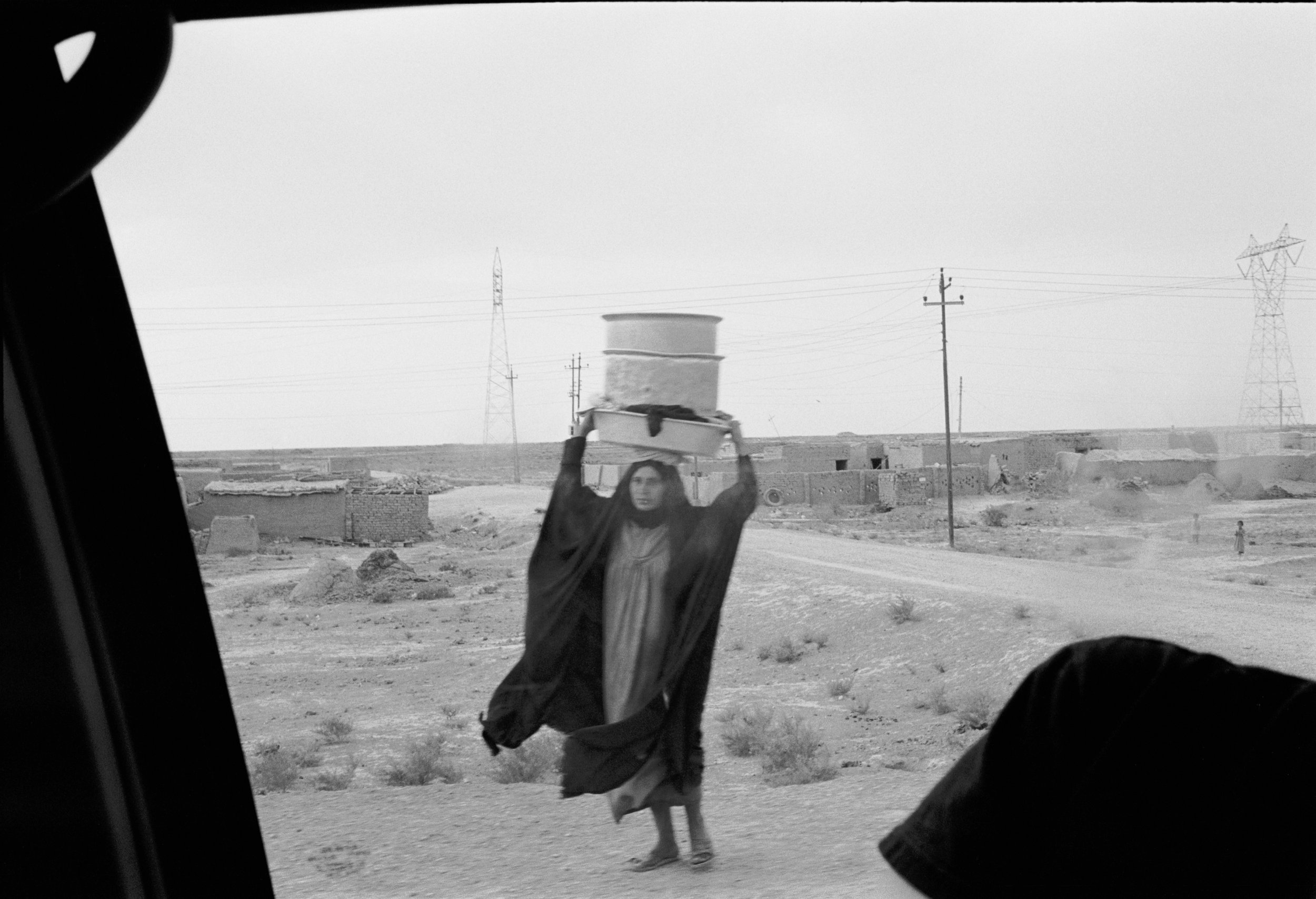  A woman walks along the highway going from Baghdad to Basra carrying a stack of pots. 