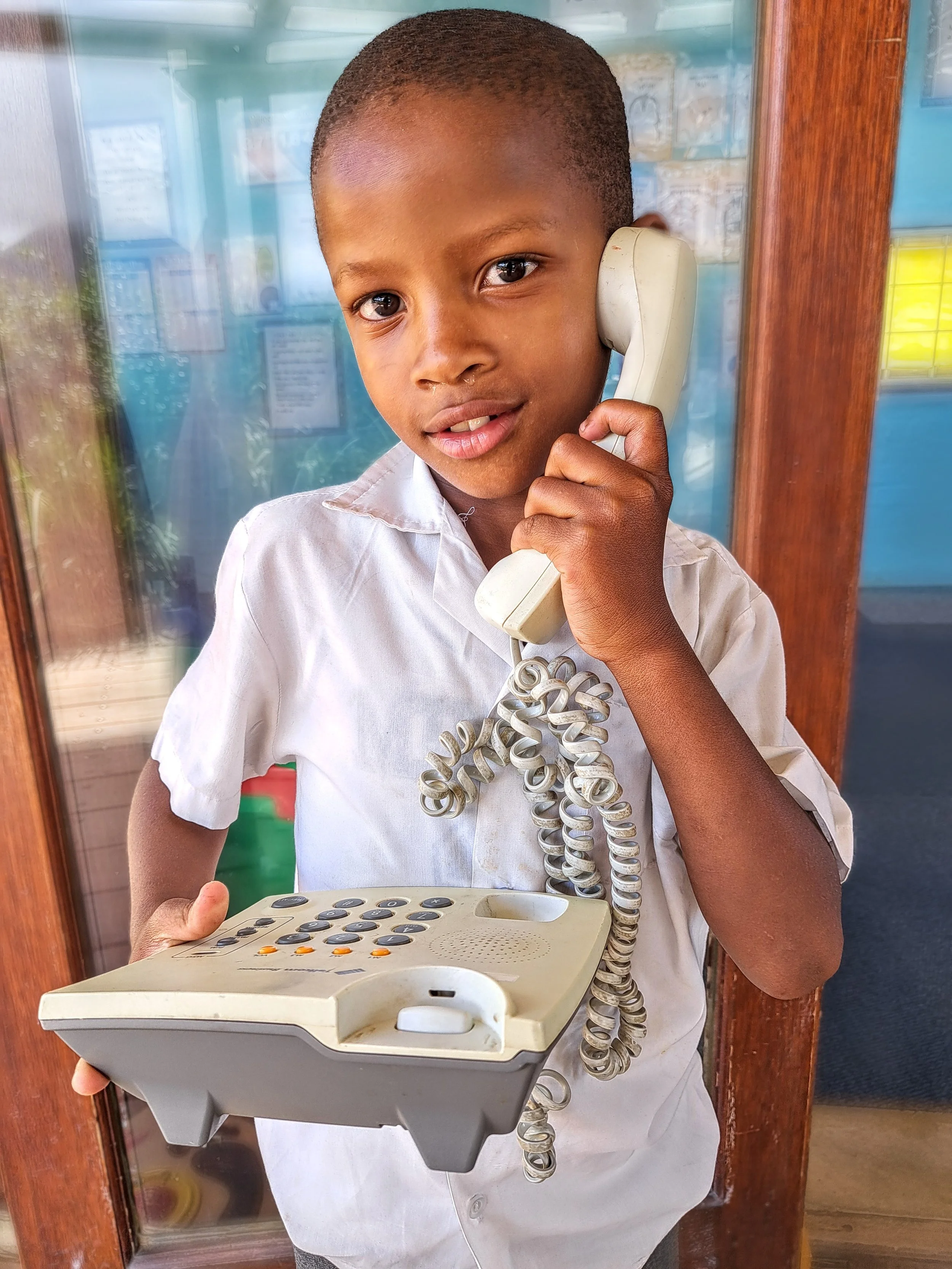 A young boy holds the receiver of a phone to his ear and smiles at the camera