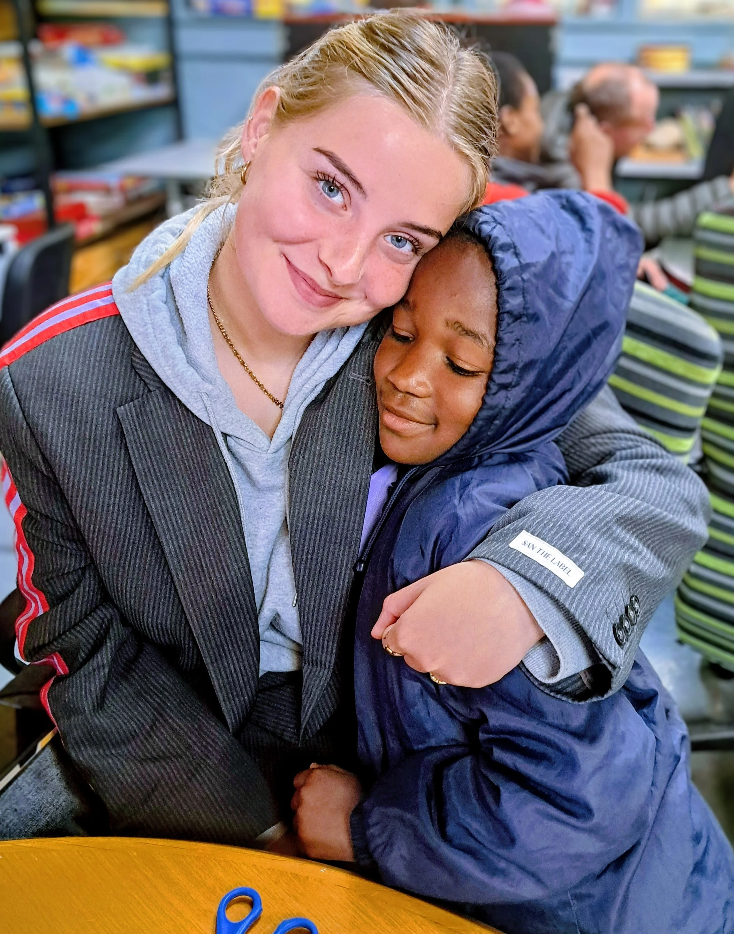 A young female volunteer hugs a child while sat at a table doing homework with them.