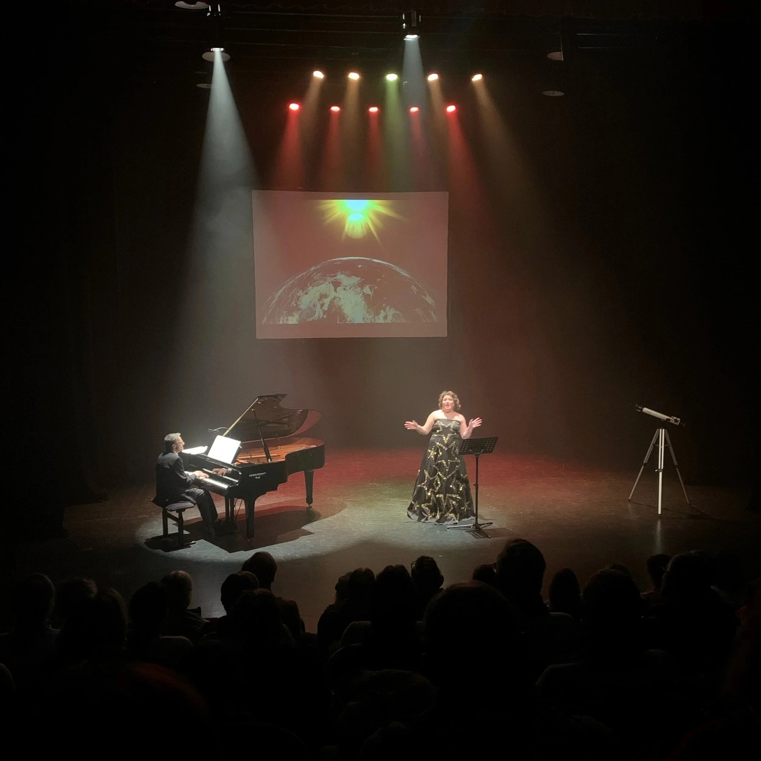 A live performance on stage with a woman singing at a microphone, a man playing the piano, a large projection of Earth's moon and the sun in the background, and a telescope to the right. Audience members are visible in the foreground.