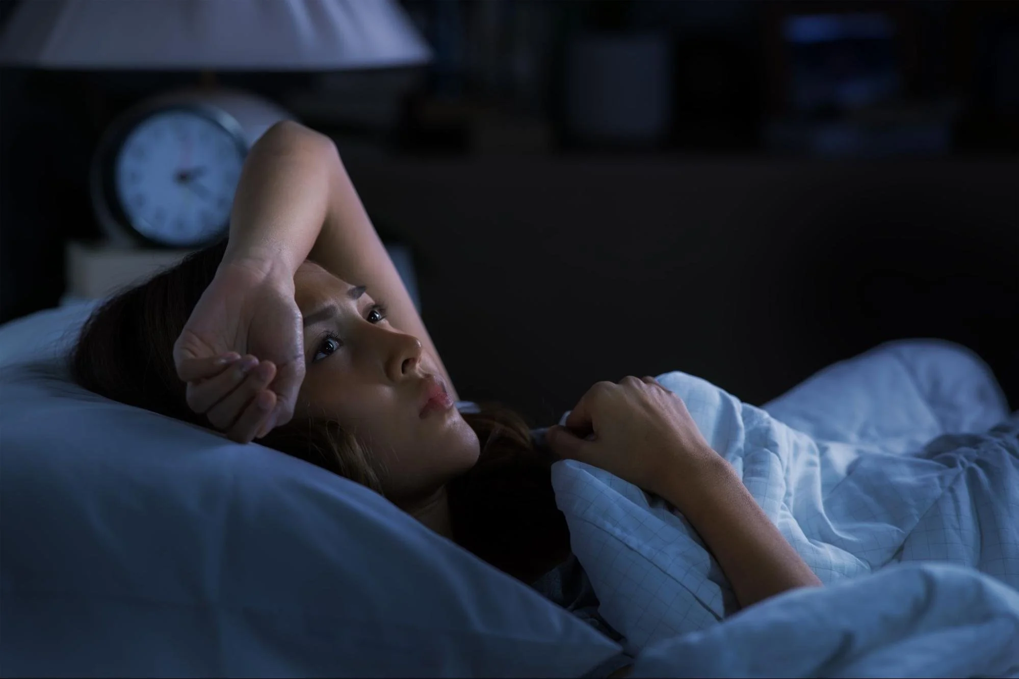 An anxious woman lying awake in bed in a dark room, looking at the ceiling near a nightstand clock.