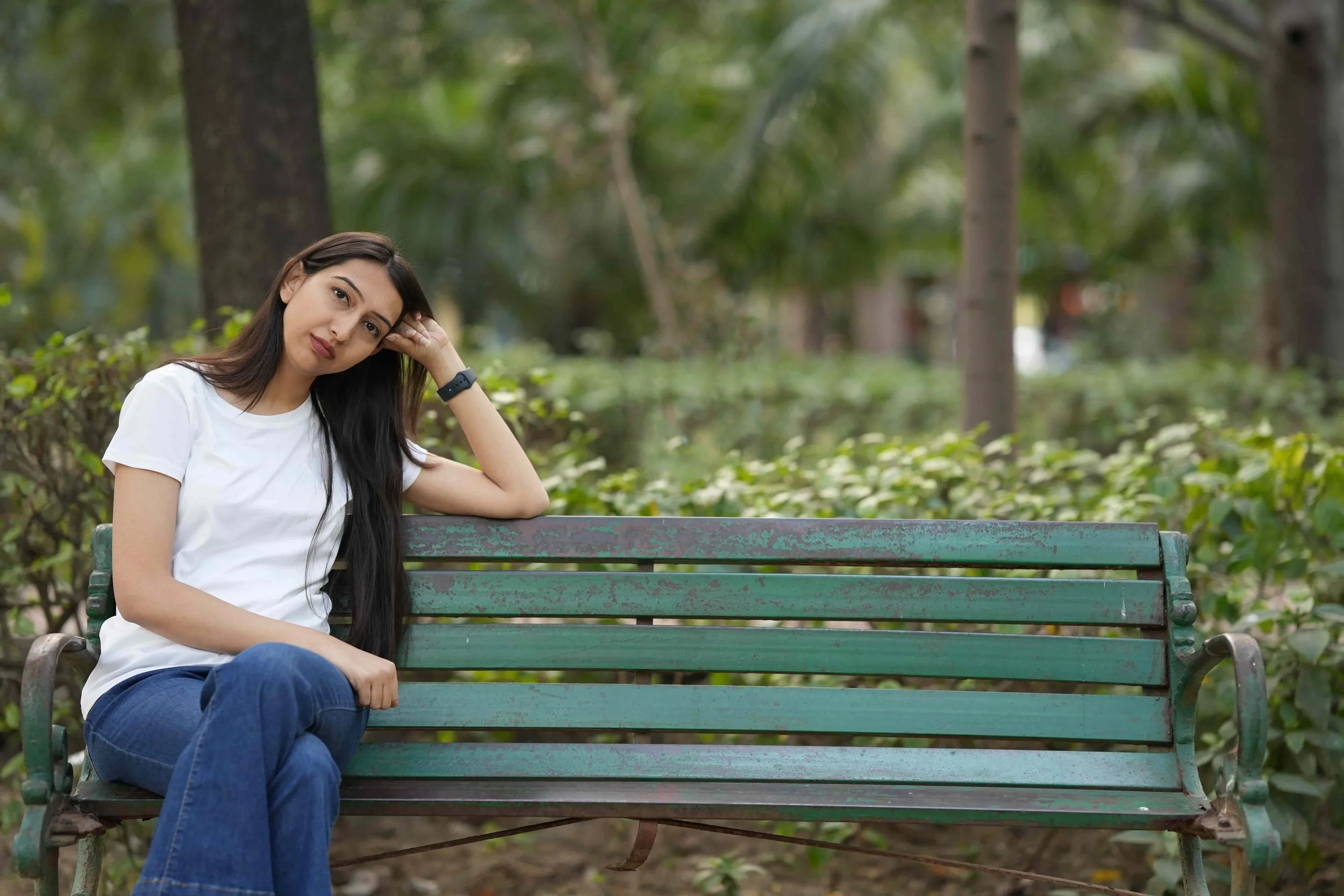 A pensive young woman sitting alone on a park bench resting her head on her hand.