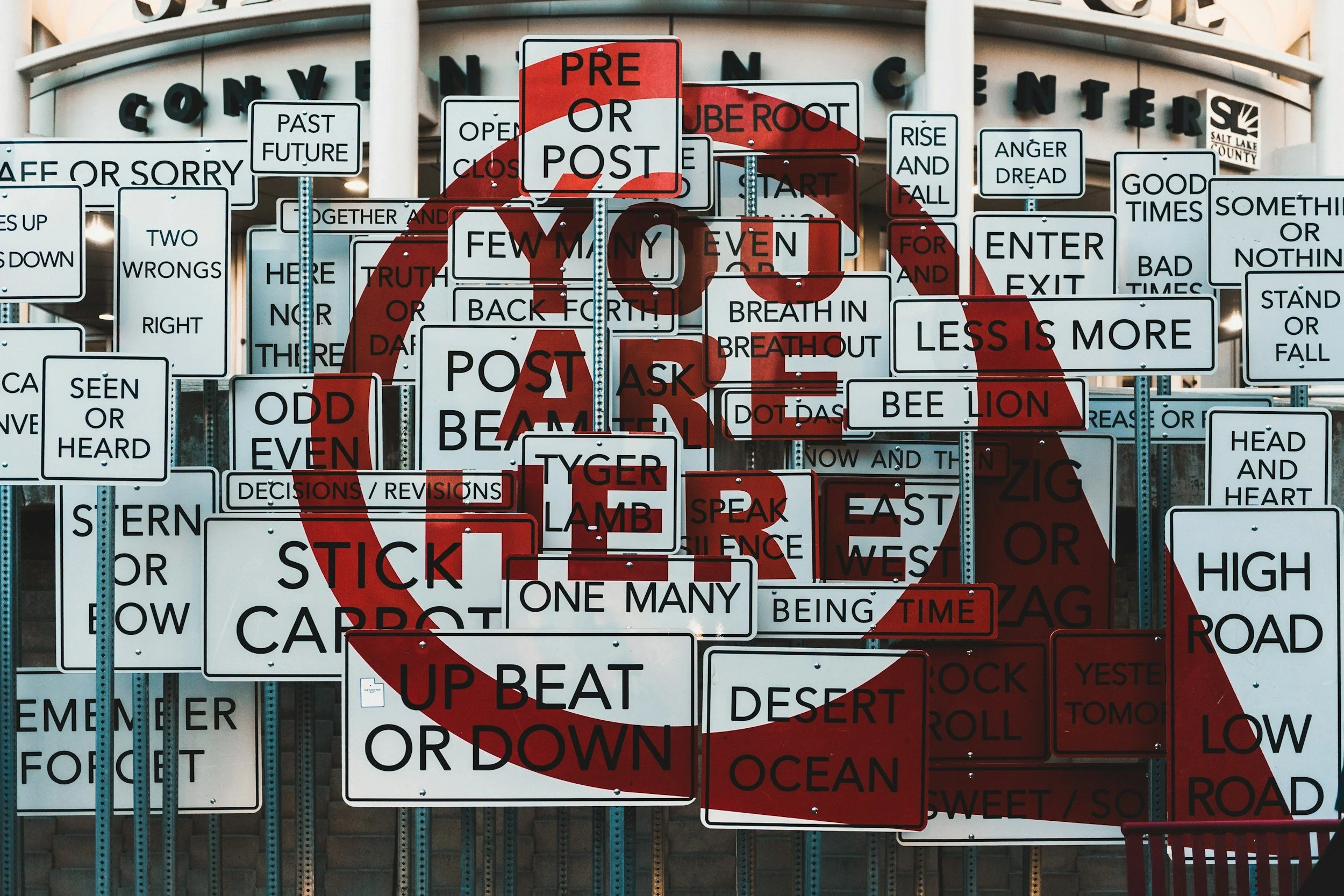 a huge number of white street signs blocks a convention center. They read things like "stick carrot" "pre or post" "rise and fall". Over top, and huge red circle contains the words YOU ARE HERE. A sort of arrow descends from the circle.