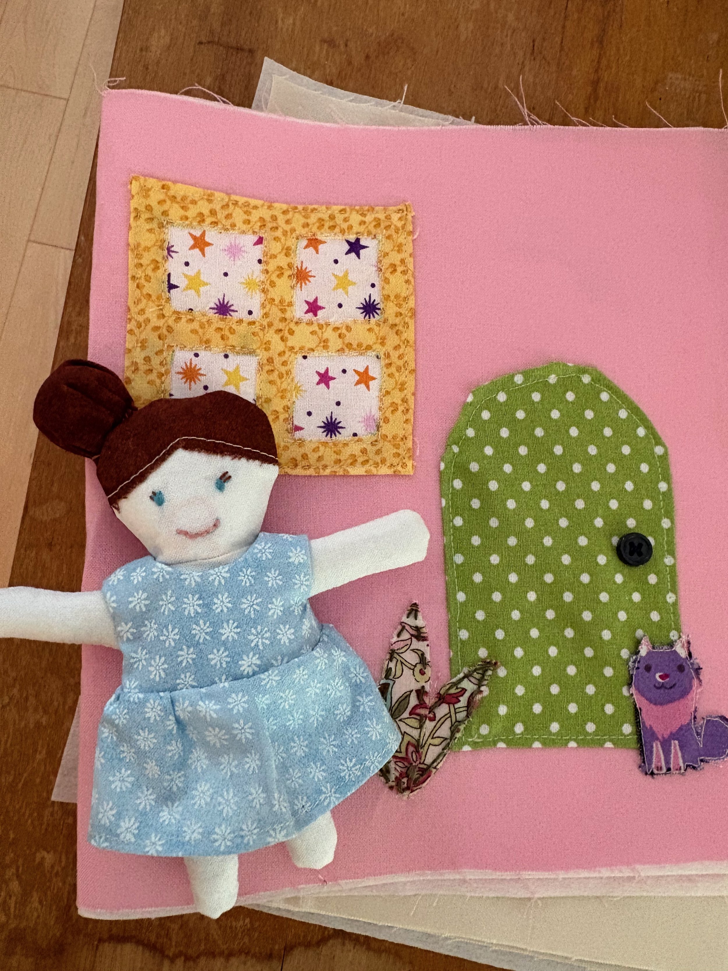 A little fabric doll with brown hair and a little blue dress in front of a pink cover with a door, a window, a plant and a purple cat