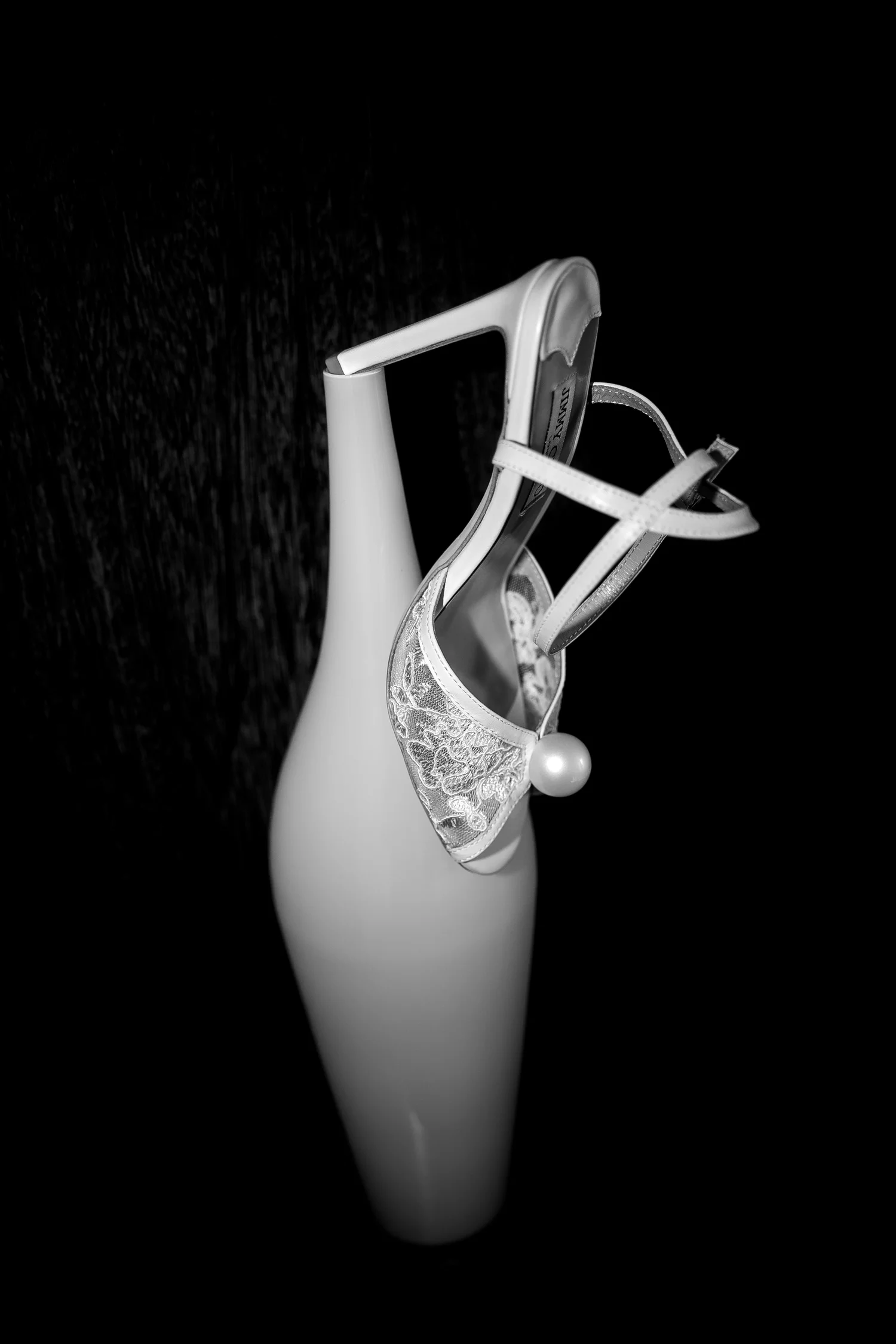 Black-and-white close-up of a lace bridal sandal with a pearl detail, balanced on a white vase