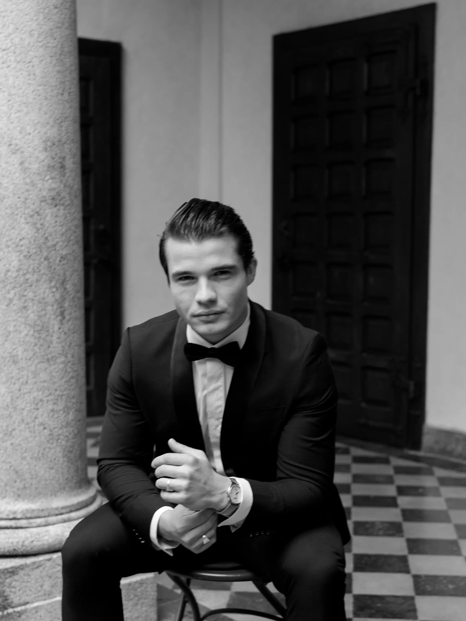 Black-and-white portrait of a groom in a tuxedo seated beside a stone column
