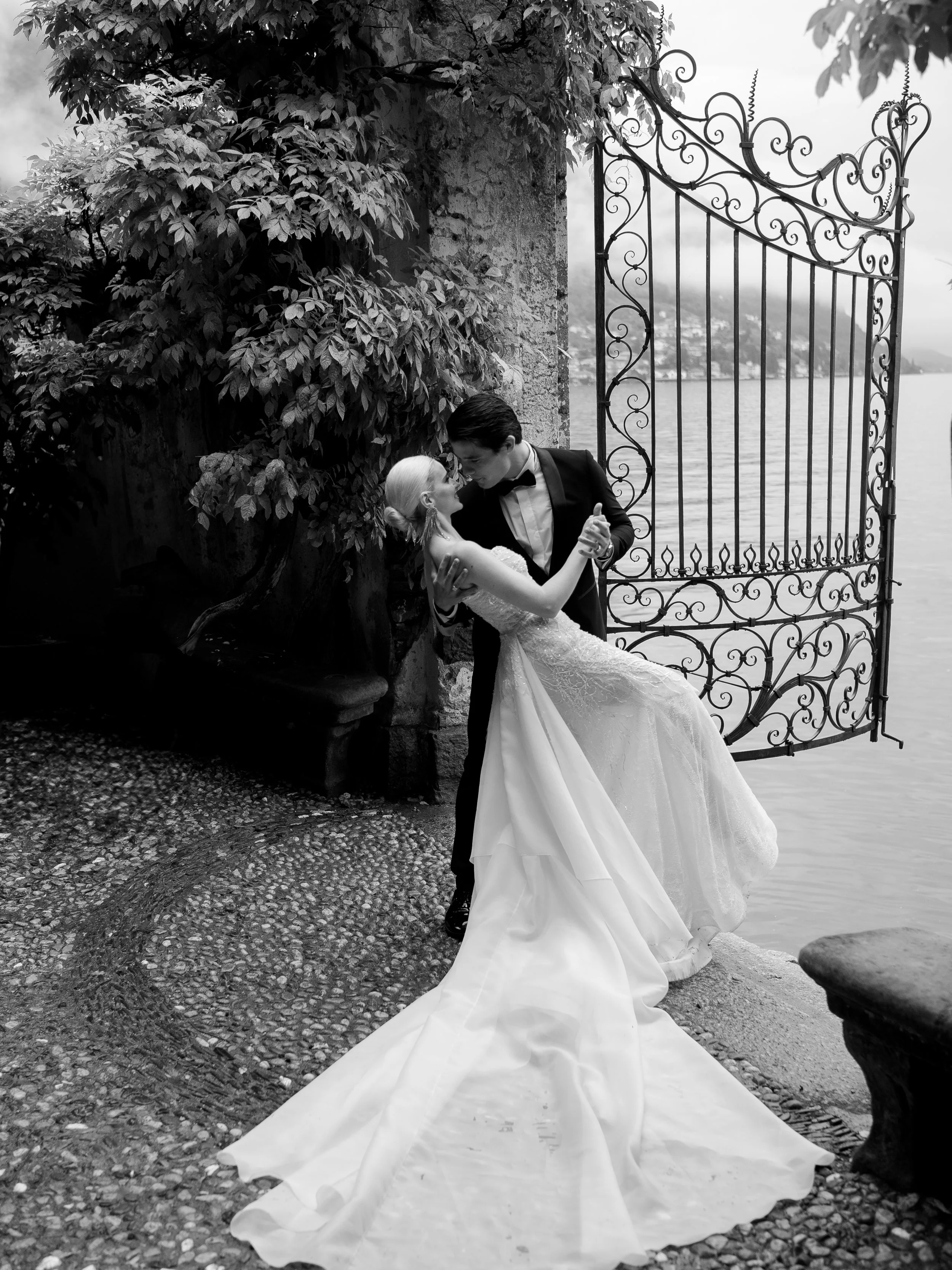 A wedding couple dancing outdoors near a body of water, with a decorative wrought iron gate and lush greenery, in a black-and-white photo.