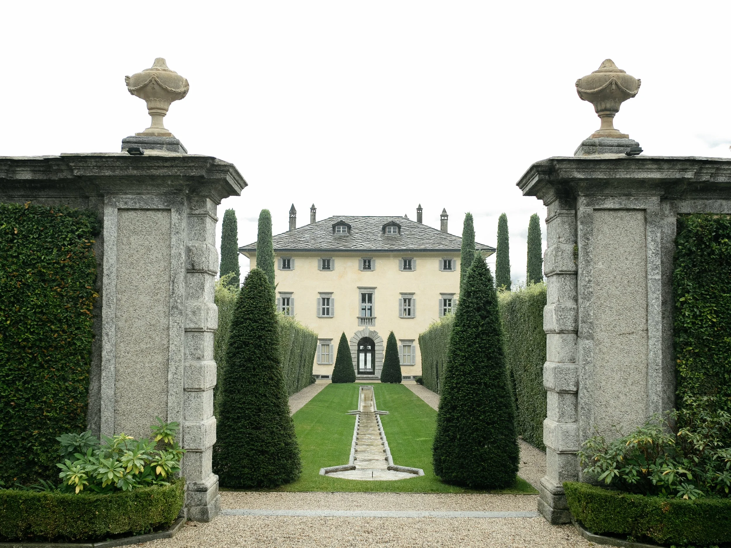 View of a formal garden with a central water feature leading to a large, elegant white mansion framed by stone pillars topped with urn-shaped ornaments, surrounded by tall, manicured trees and hedges.