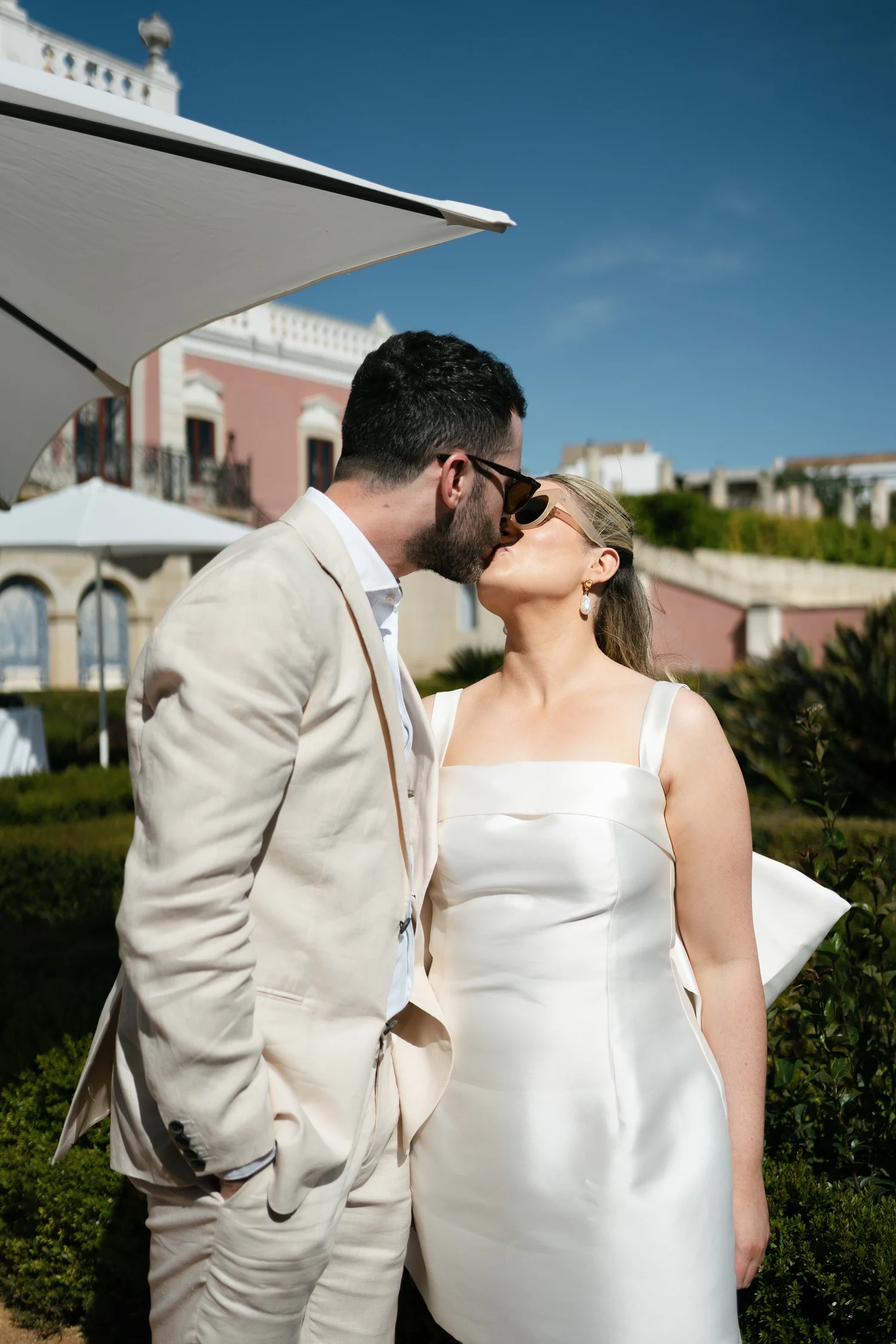 Newlyweds wearing sunglasses and sharing a kiss outside a pink wedding venue