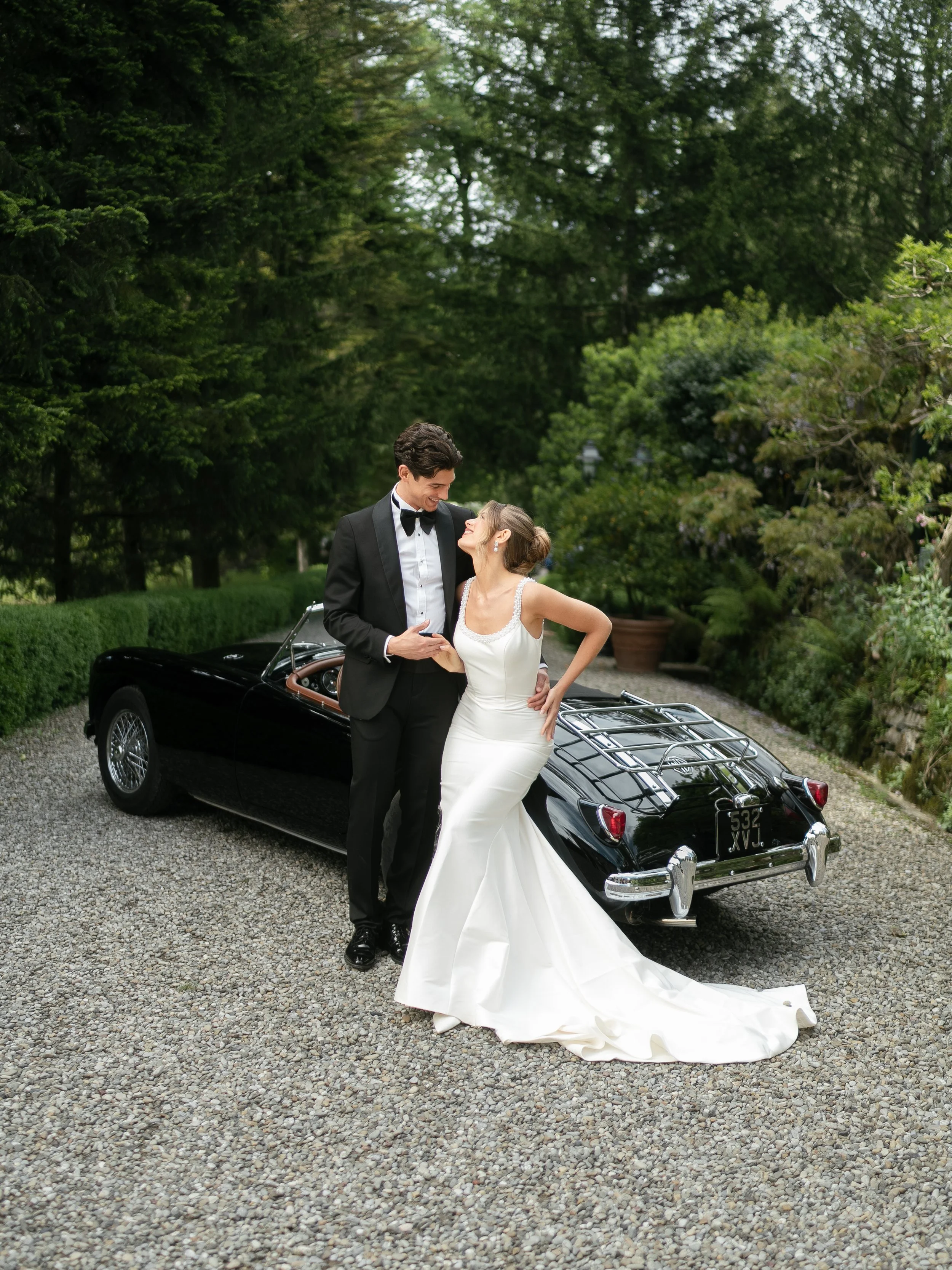 A bride and groom in wedding attire standing next to a vintage black convertible car on a gravel driveway surrounded by lush green trees and bushes.