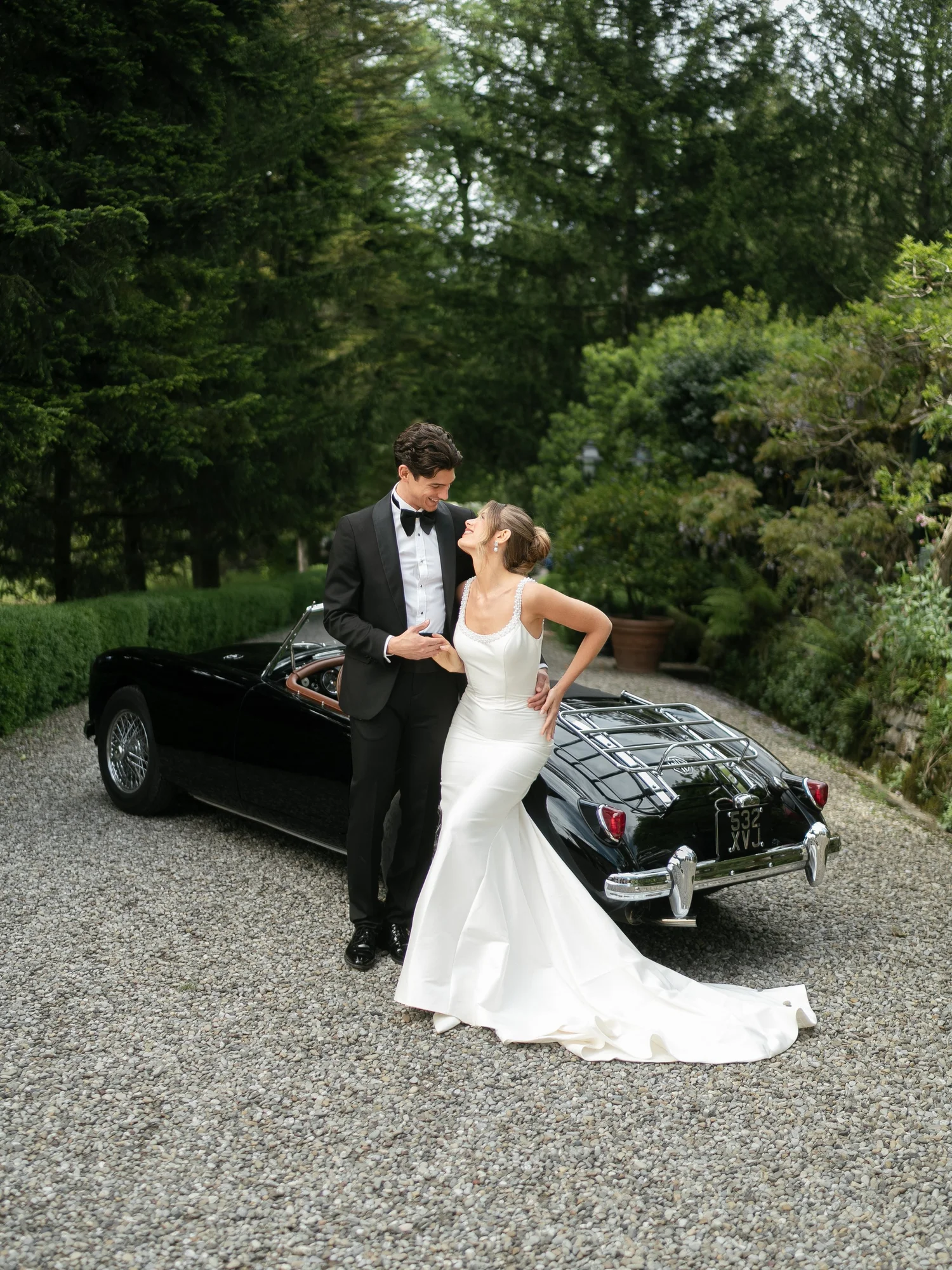 Bride and groom laughing beside a black vintage convertible on a garden driveway