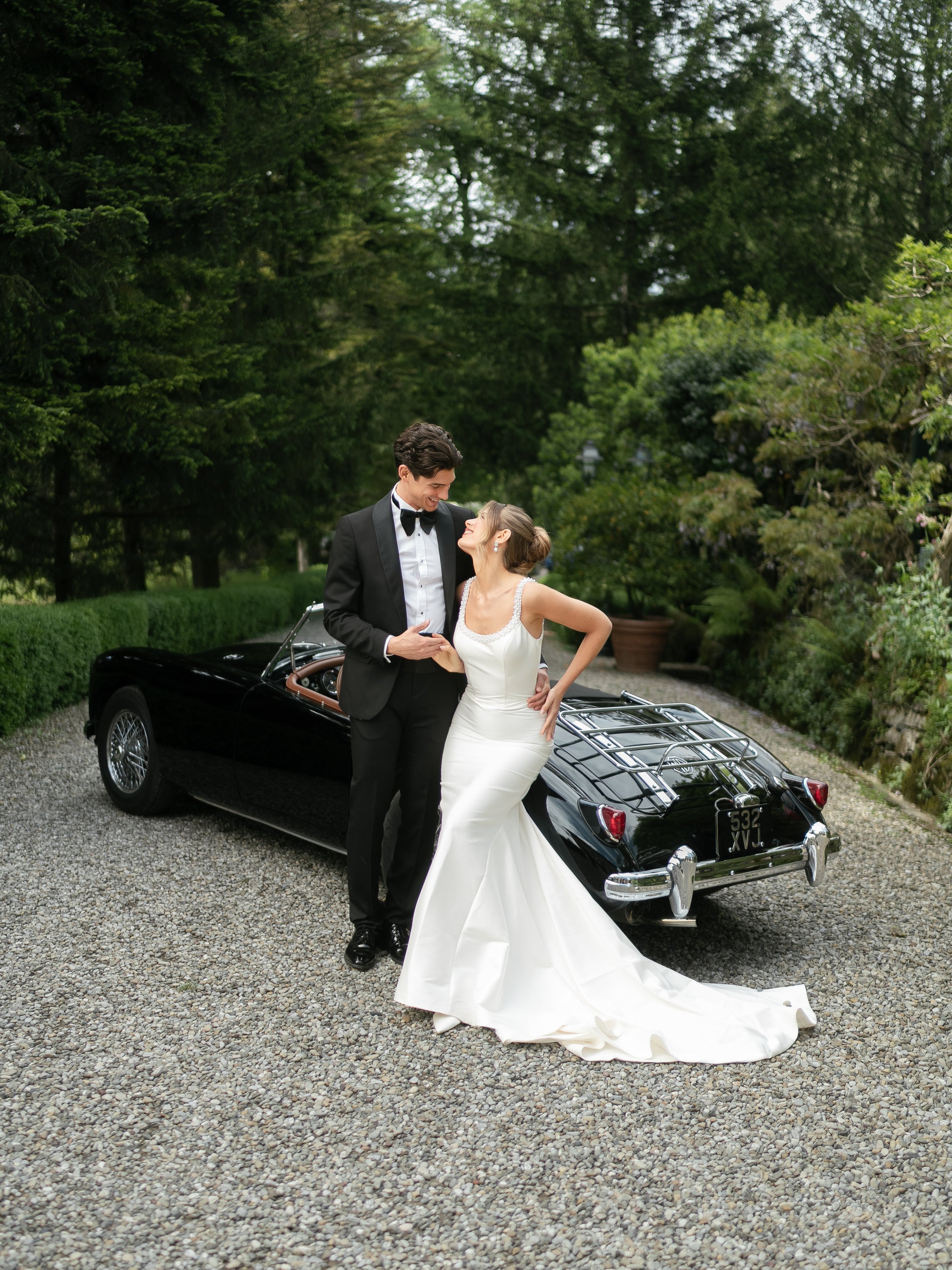 A bride and groom in wedding attire standing beside a black vintage car on a gravel driveway, surrounded by lush greenery.