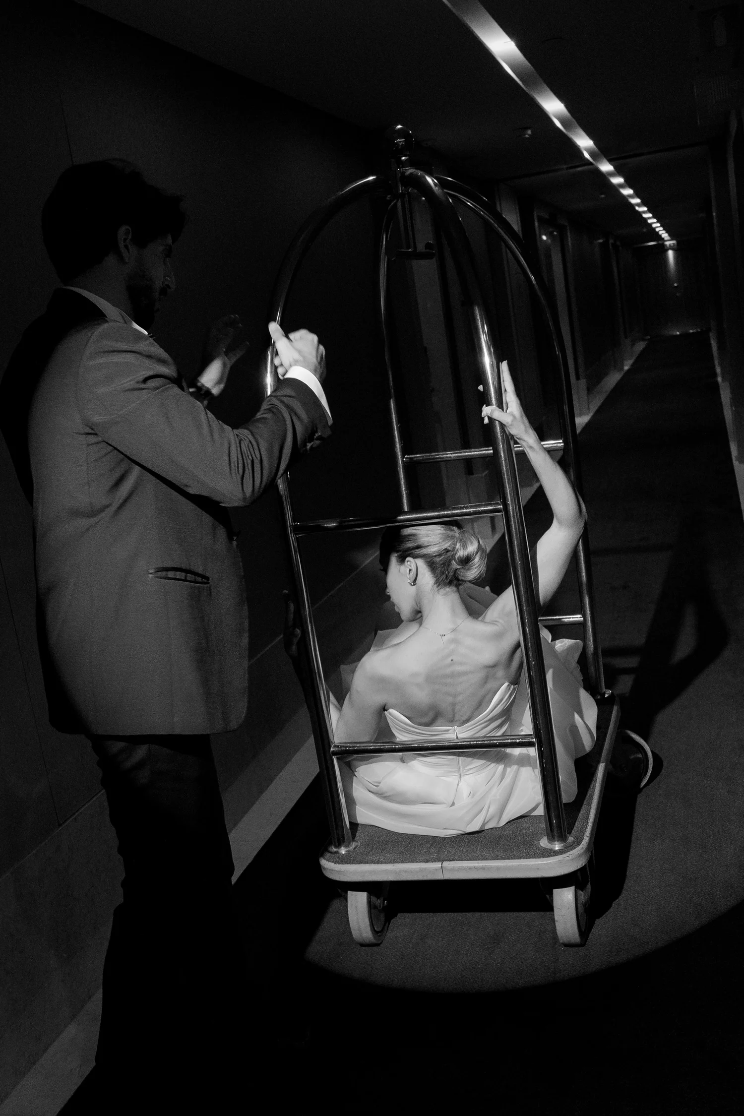 Playful black-and-white wedding portrait of a bride riding a luggage trolley with her groom