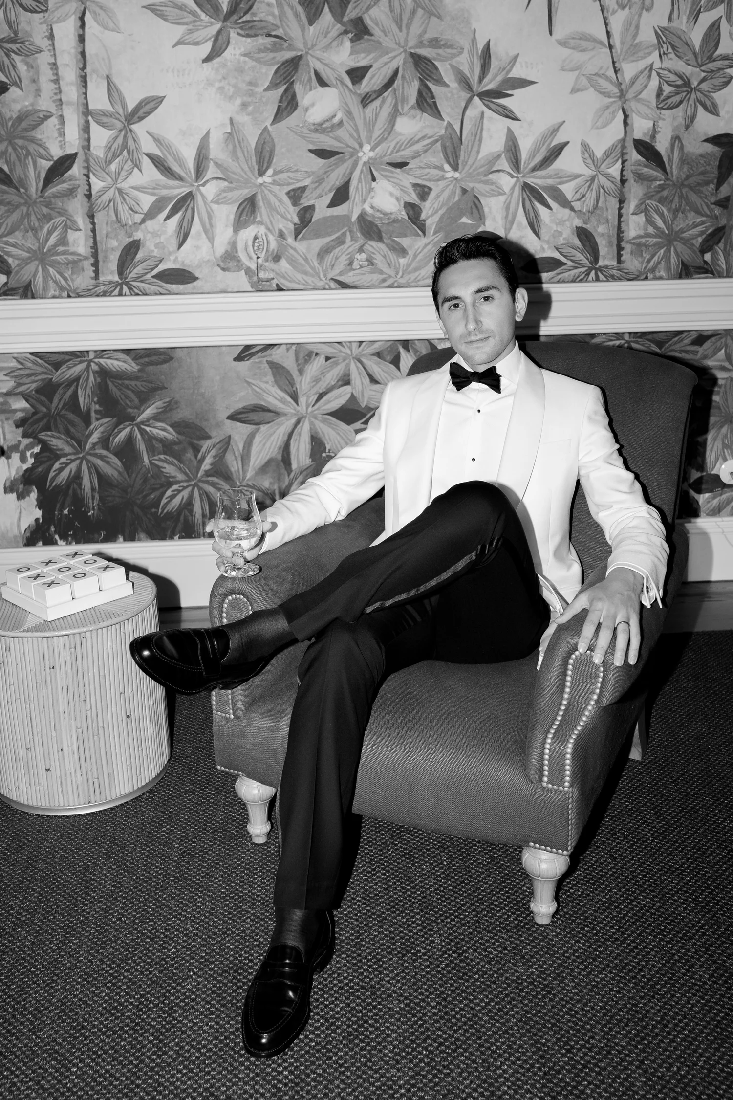 Black-and-white portrait of a groom in an ivory tuxedo seated in an armchair with a drink